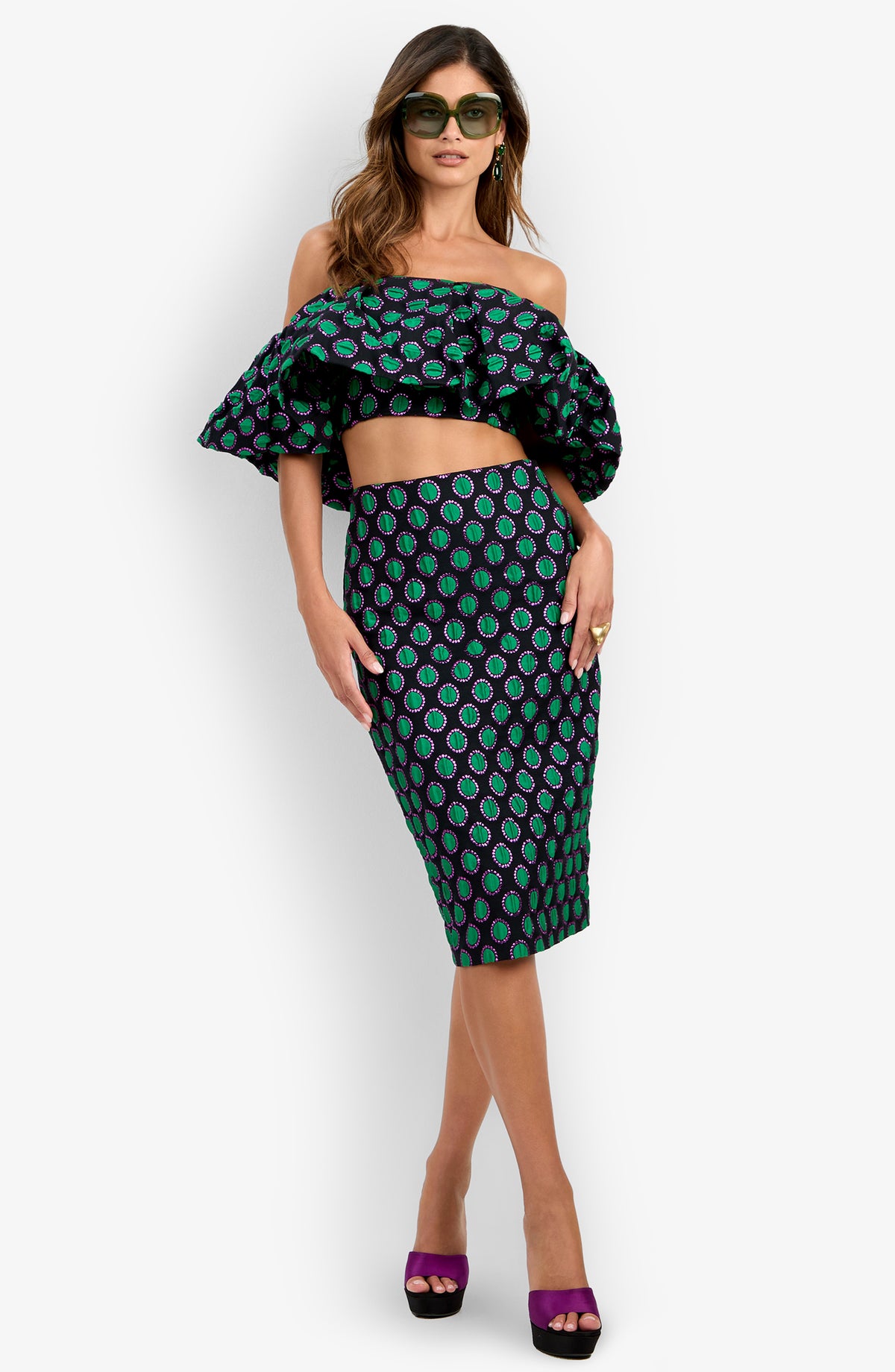 Woman in sunglasses wearing the Dafinas Top—an off-shoulder ruffled crop top with a green and purple pattern—paired with a matching pencil skirt and purple open-toe heels, posing against a plain white background.