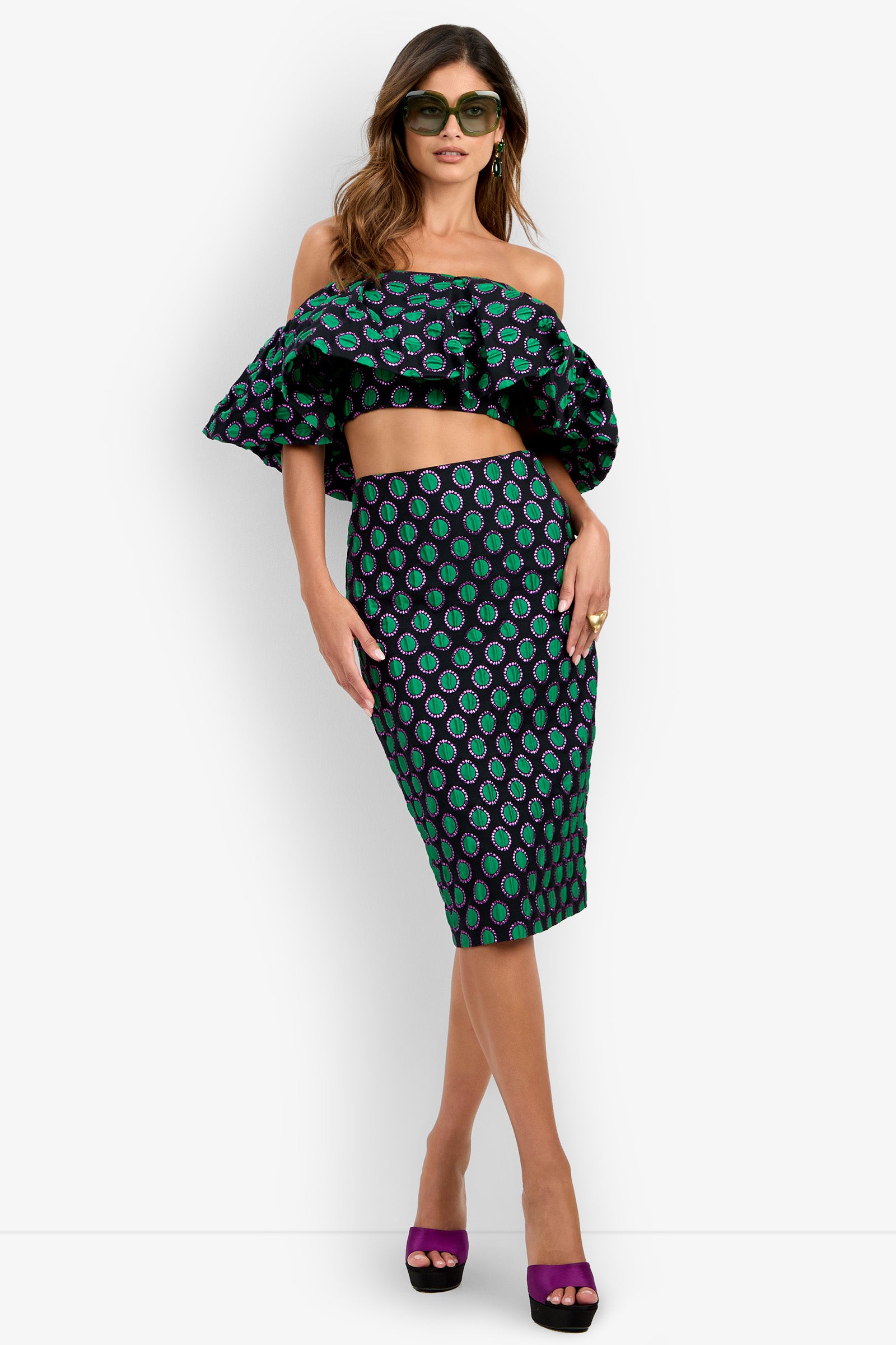 Wearing chic sunglasses, she exudes confidence in the Dafinas Top—a ruffled off-shoulder crop top—paired with a matching high-waisted pencil skirt featuring a striking green and purple pattern. Purple open-toe heels complete her look against a plain background.