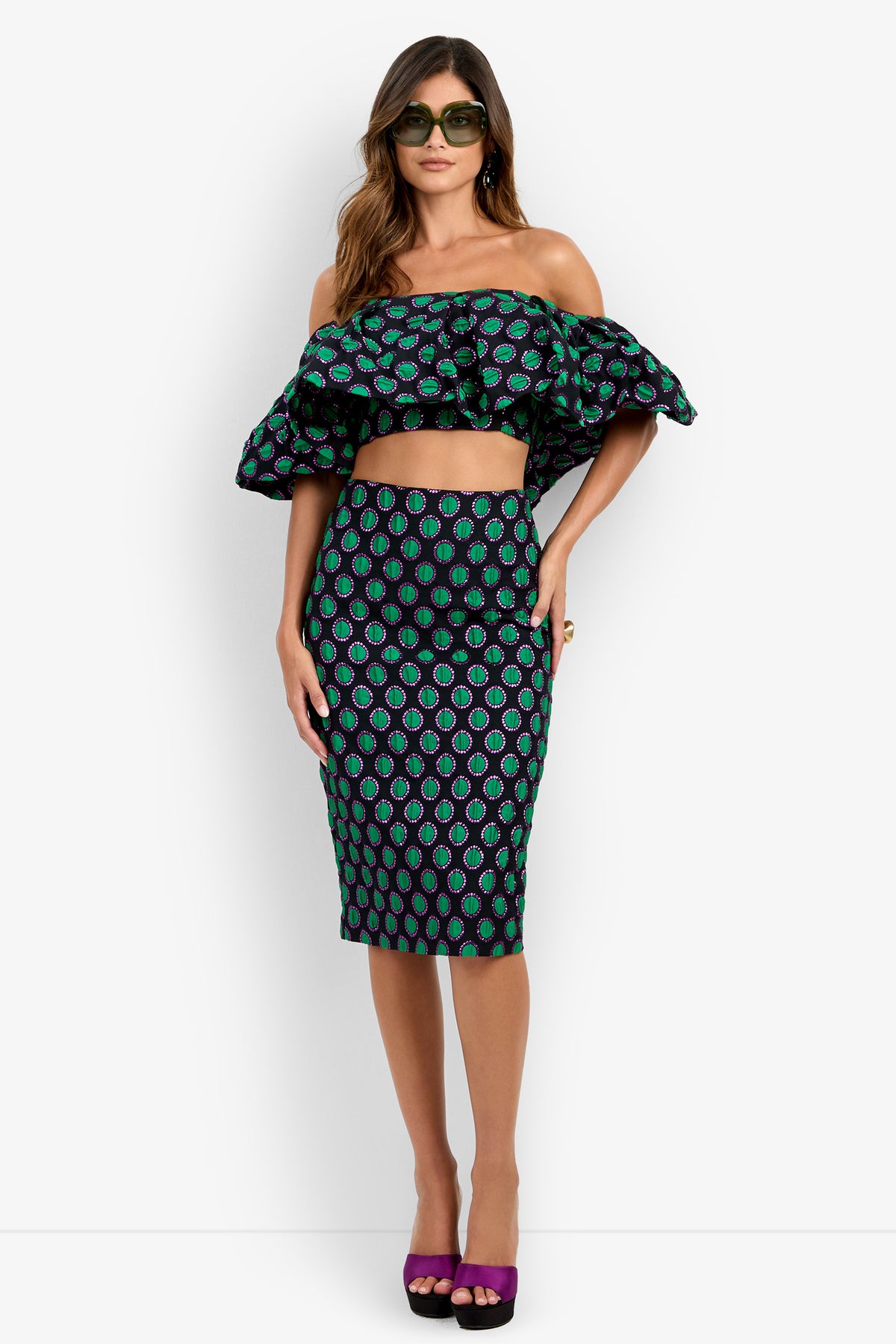 A woman wearing sunglasses, an off-shoulder ruffled crop top, and the Soniah Skirt—a high-waisted pencil skirt featuring a black, green, and purple patterned print—stands against a white background.