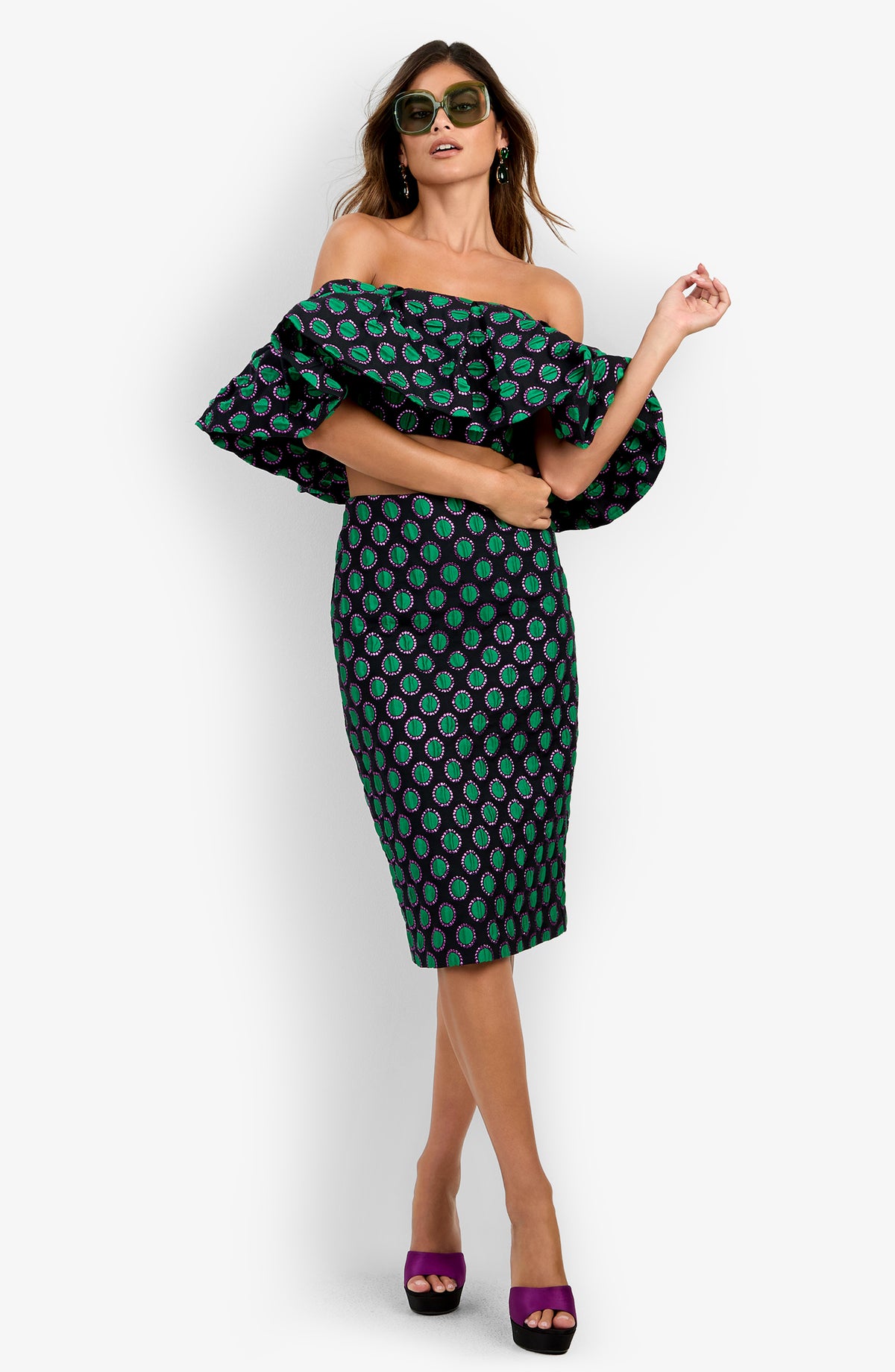 A woman wearing the Dafinas Top, featuring green and purple patterns, paired with sunglasses and purple high heels, poses against a white background.