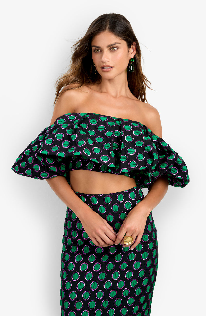 A woman wearing the Dafinas Top—an off-the-shoulder crop top featuring a green and purple circular pattern on black fabric—paired with a matching skirt, poses with her hands clasped.