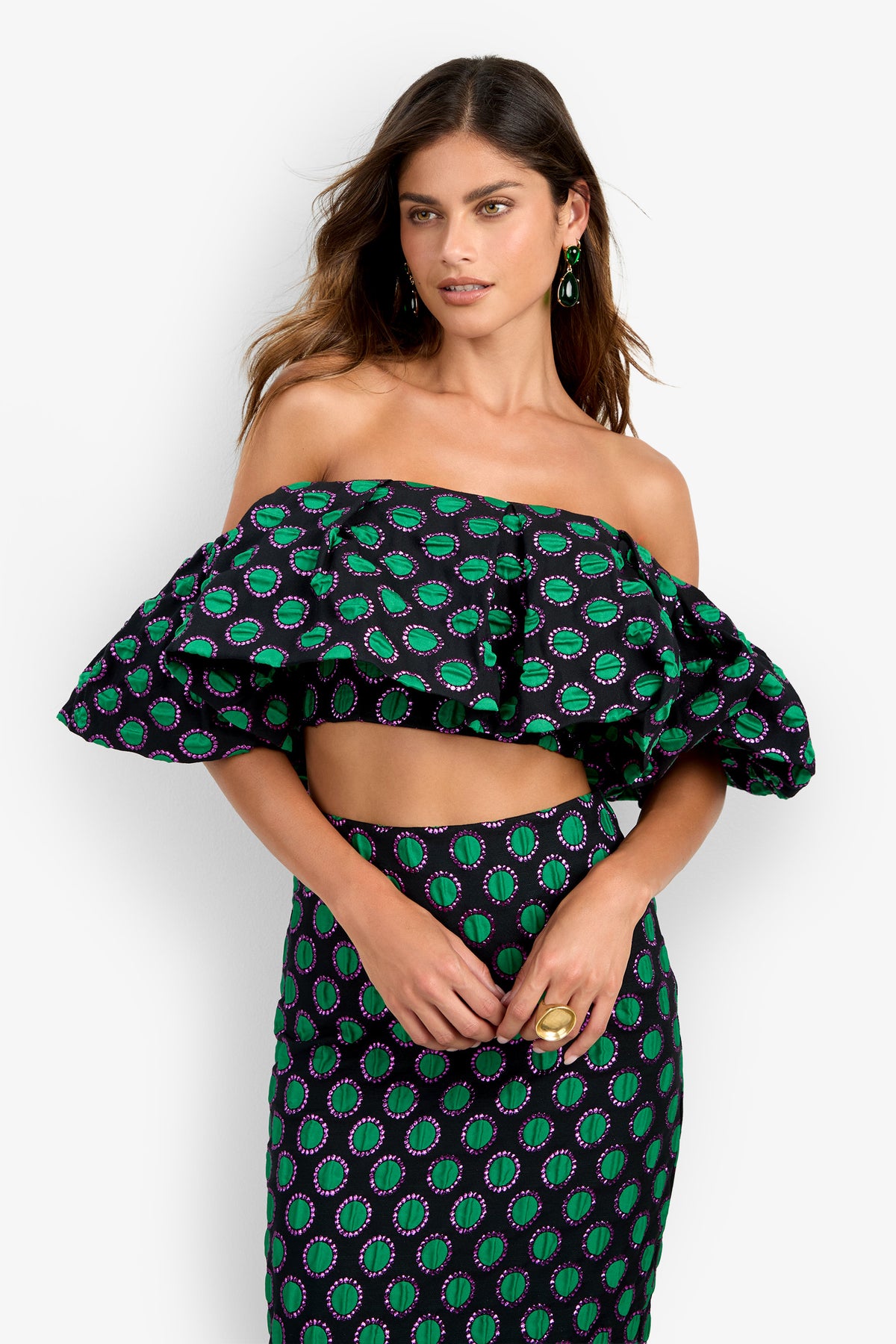 A woman wearing the Dafinas Top—an off-shoulder, ruffled crop top featuring a green and purple geometric pattern on a black background—paired with a matching high-waisted skirt. She has long brown hair and accessorizes with earrings and a ring.