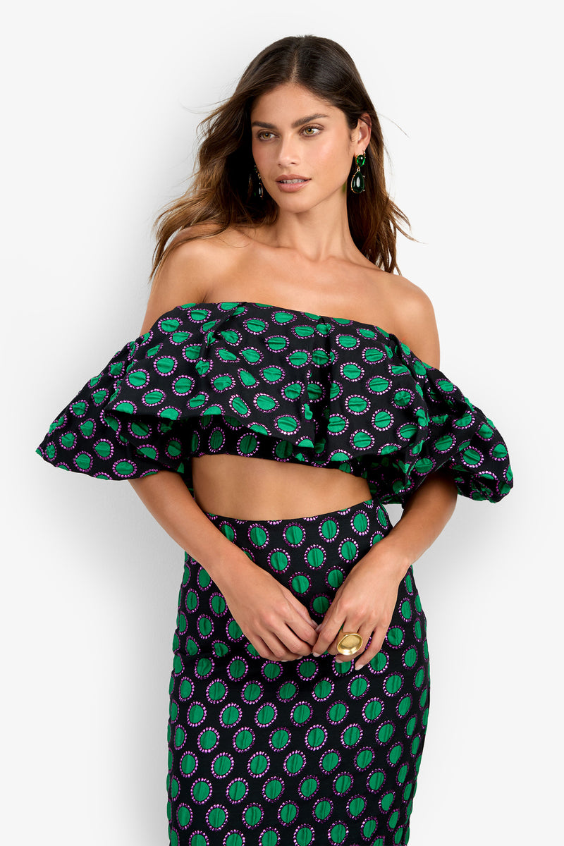 A woman wearing the Dafinas Top—an off-shoulder, ruffled crop top featuring a green and purple geometric pattern on a black background—paired with a matching high-waisted skirt. She has long brown hair and accessorizes with earrings and a ring.