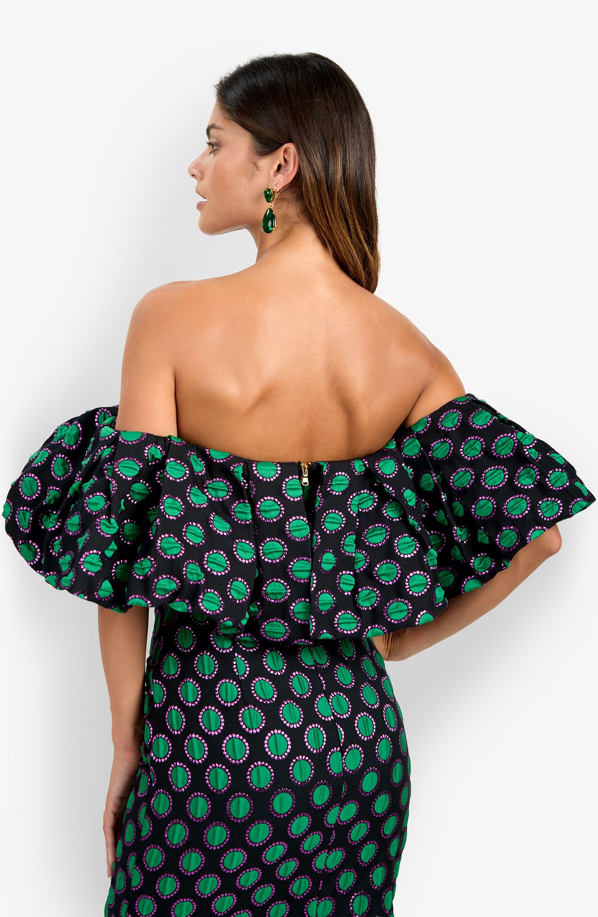 Woman facing away, wearing the Dafinas Top featuring an off-the-shoulder design with ruffled sleeves and a green and purple geometric pattern. She has long brown hair and wears green earrings.