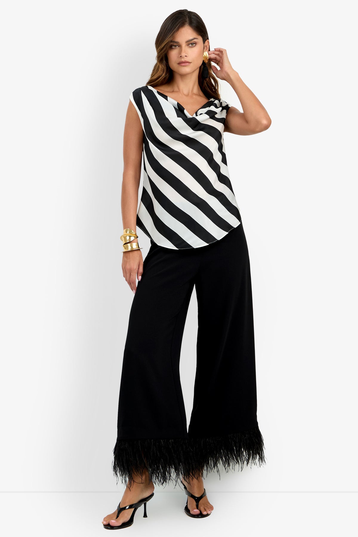 Woman wearing the Alessi Top, a black and white striped sleeveless top, paired with wide-leg black pants featuring feathered hems, black sandals, and gold jewelry, standing against a plain white background.