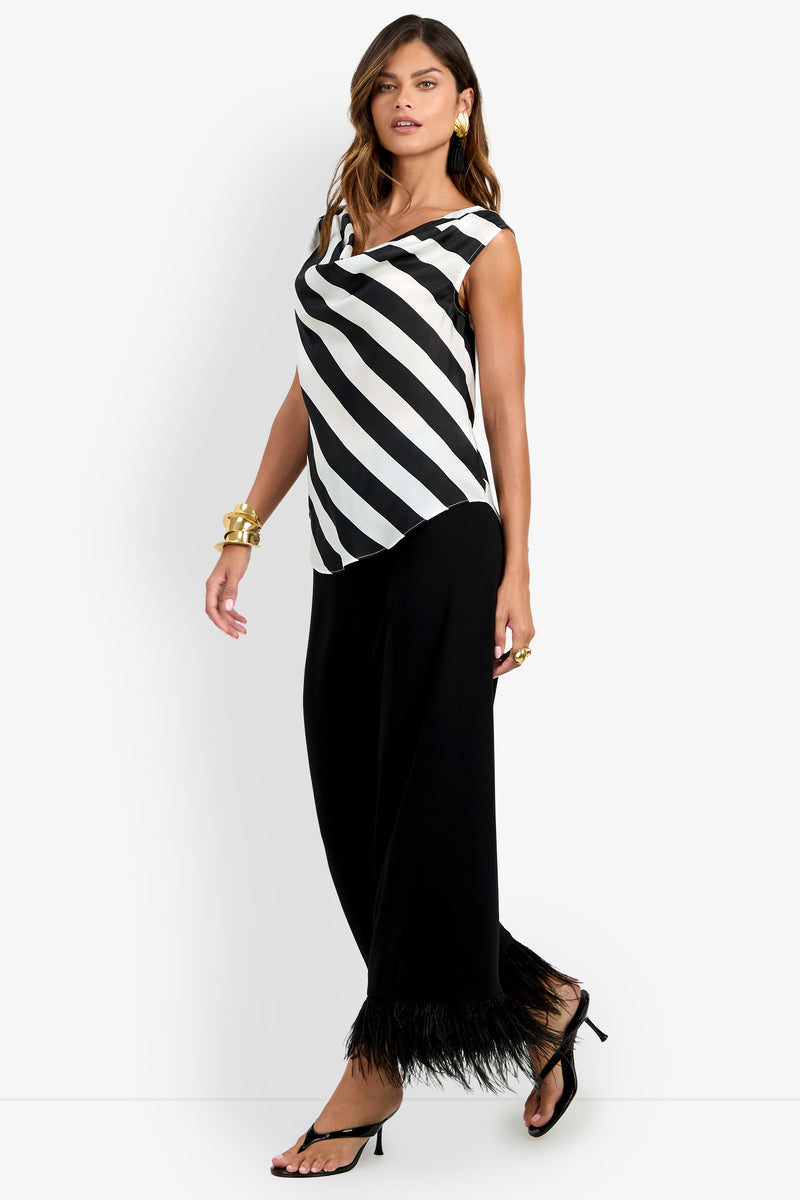A woman wearing the Alessi Top—a sleeveless black and white diagonal striped top—paired with black wide-leg pants featuring feathered hems, black heeled sandals, gold jewelry, and earrings poses against a white background.