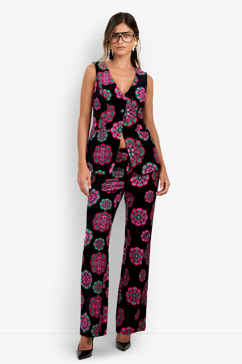 A woman wearing the Raylee Top—a sleeveless black suit adorned with bold pink, green, and blue floral patterns—paired with black heels, glasses, and earrings stands against a plain white background.