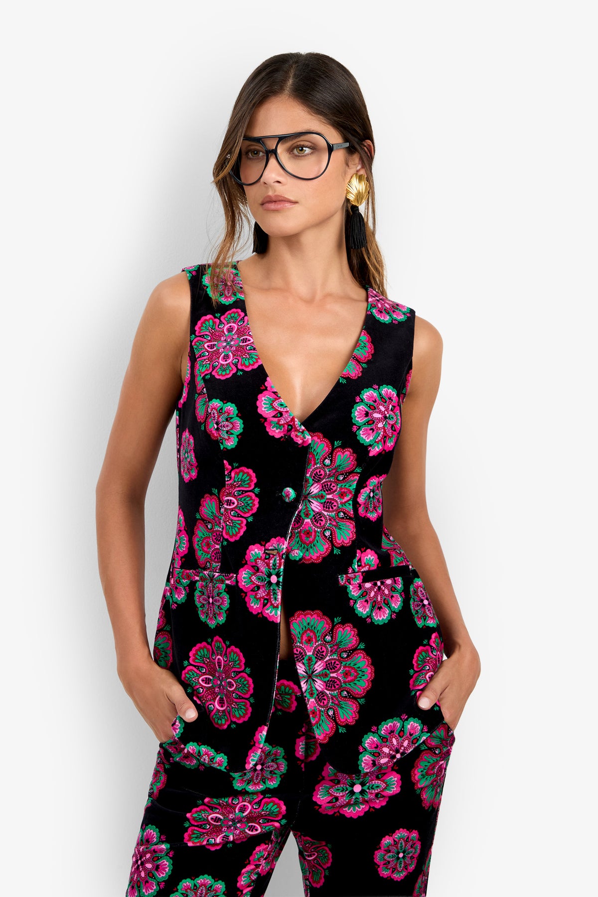 A woman stands against a white background wearing glasses and the Raylee Top—a sleeveless, black blouse adorned with bright pink and green floral patterns—paired with large gold earrings, her hands in her pockets.