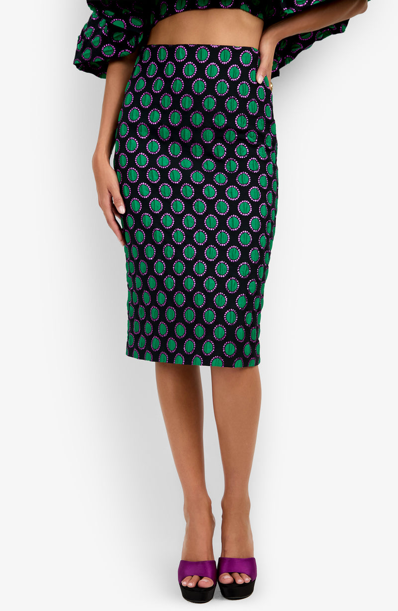 A woman wearing the Soniah Skirt, a knee-length, fitted piece featuring a green and purple circular pattern on a black background, paired with open-toe purple heels and a matching cropped top.