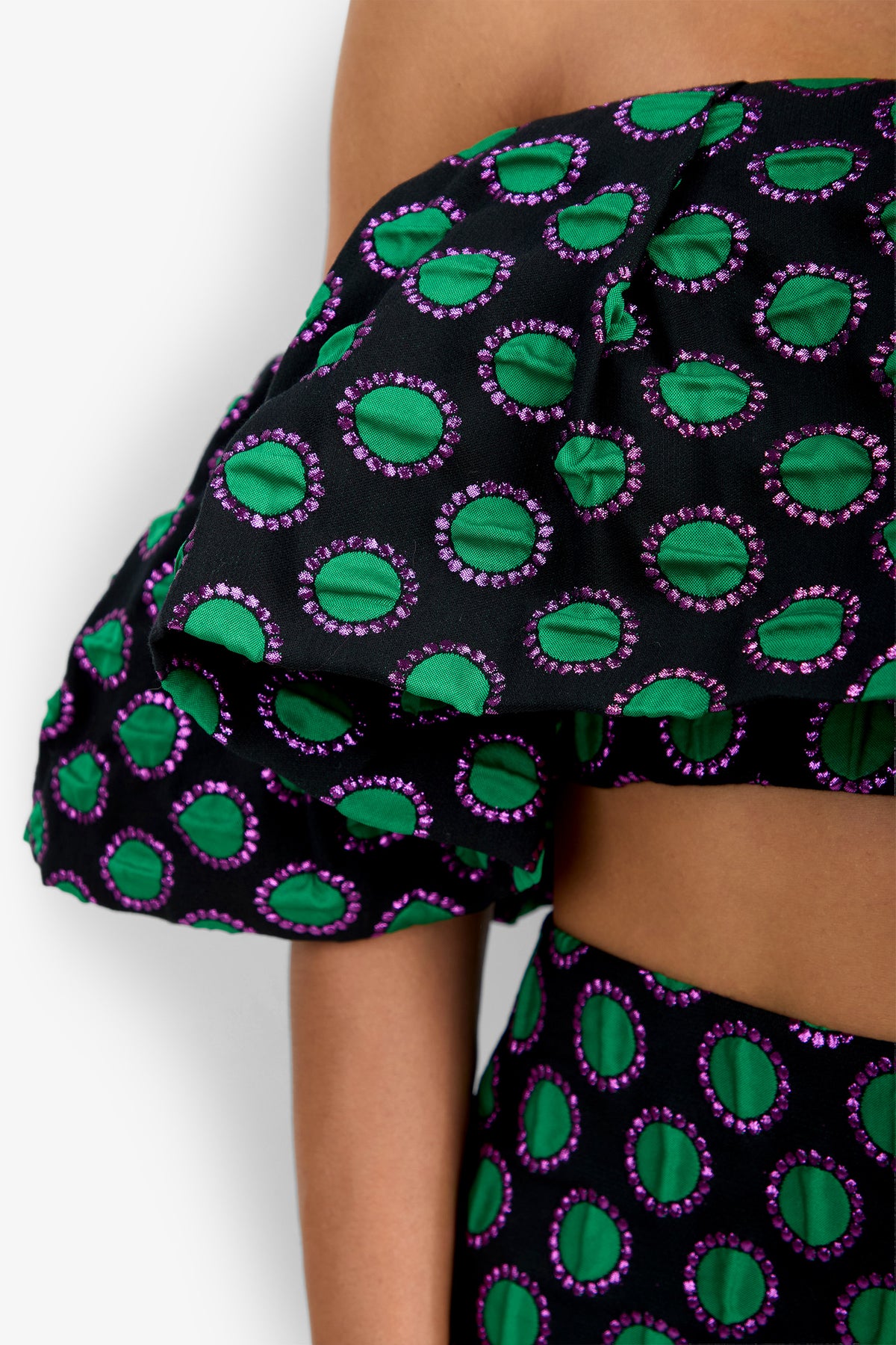 A close-up of a person wearing the Dafinas Top—an off-shoulder top crafted from black fabric adorned with green and purple circular patterns—paired with a high-waisted bottom.