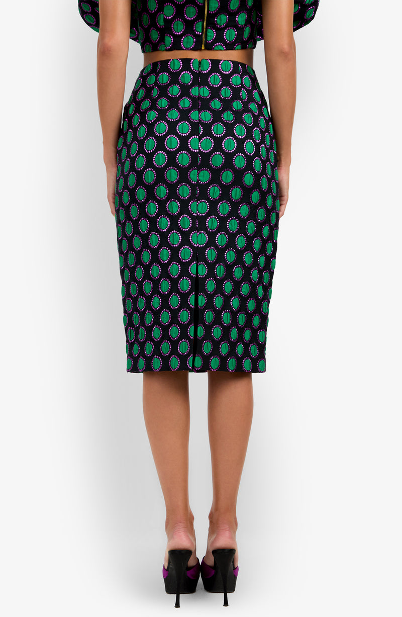 A woman stands facing away, wearing the fitted, knee-length Soniah Skirt with a green, purple, and black circular pattern. The skirt features a visible zipper down the back, and she pairs it with black high heels.