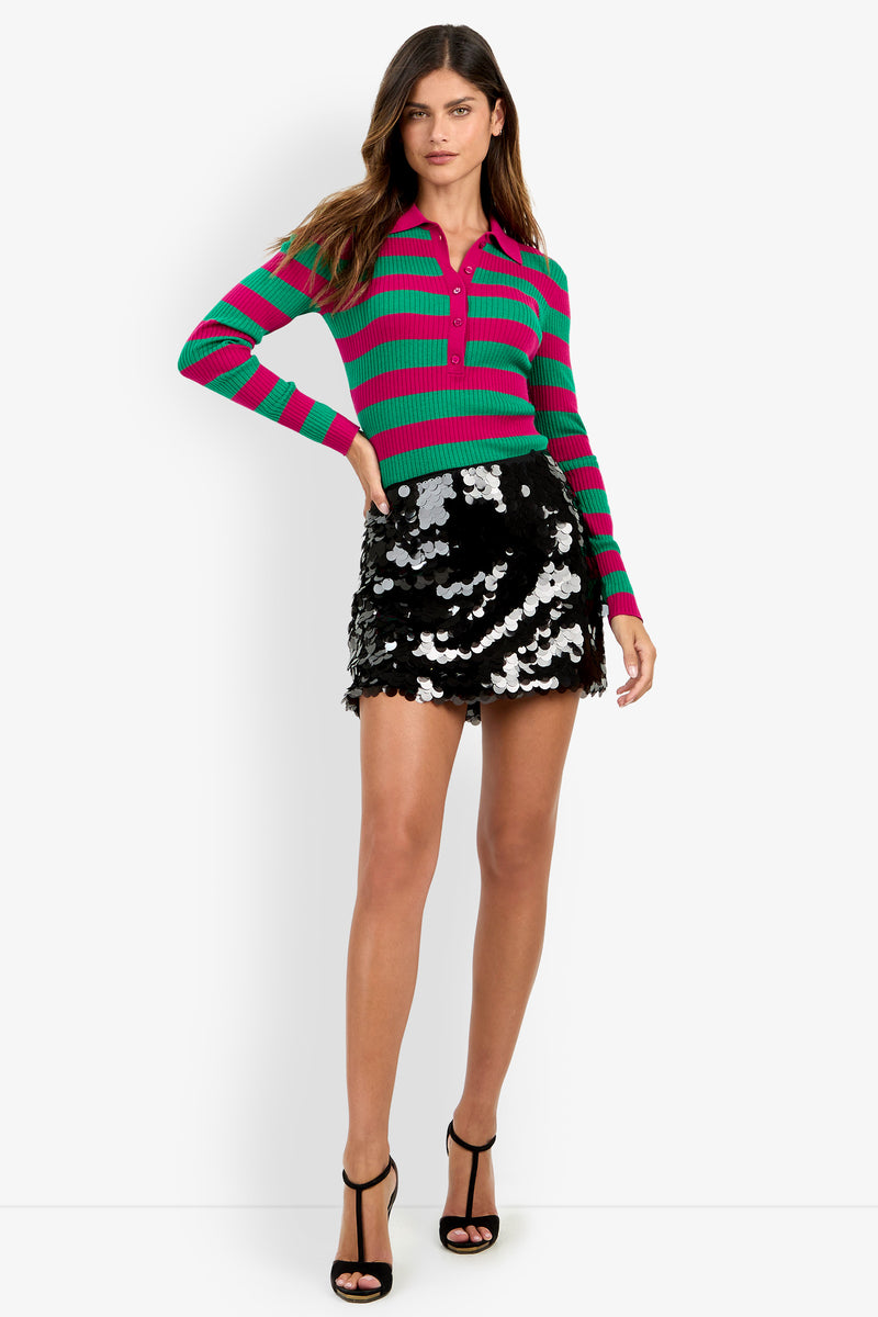 Wearing the Mini Rico Skirt in black, a woman stands against a white background, pairing it with a green and pink striped long-sleeve shirt and black high-heeled sandals adorned with large silver sequins.