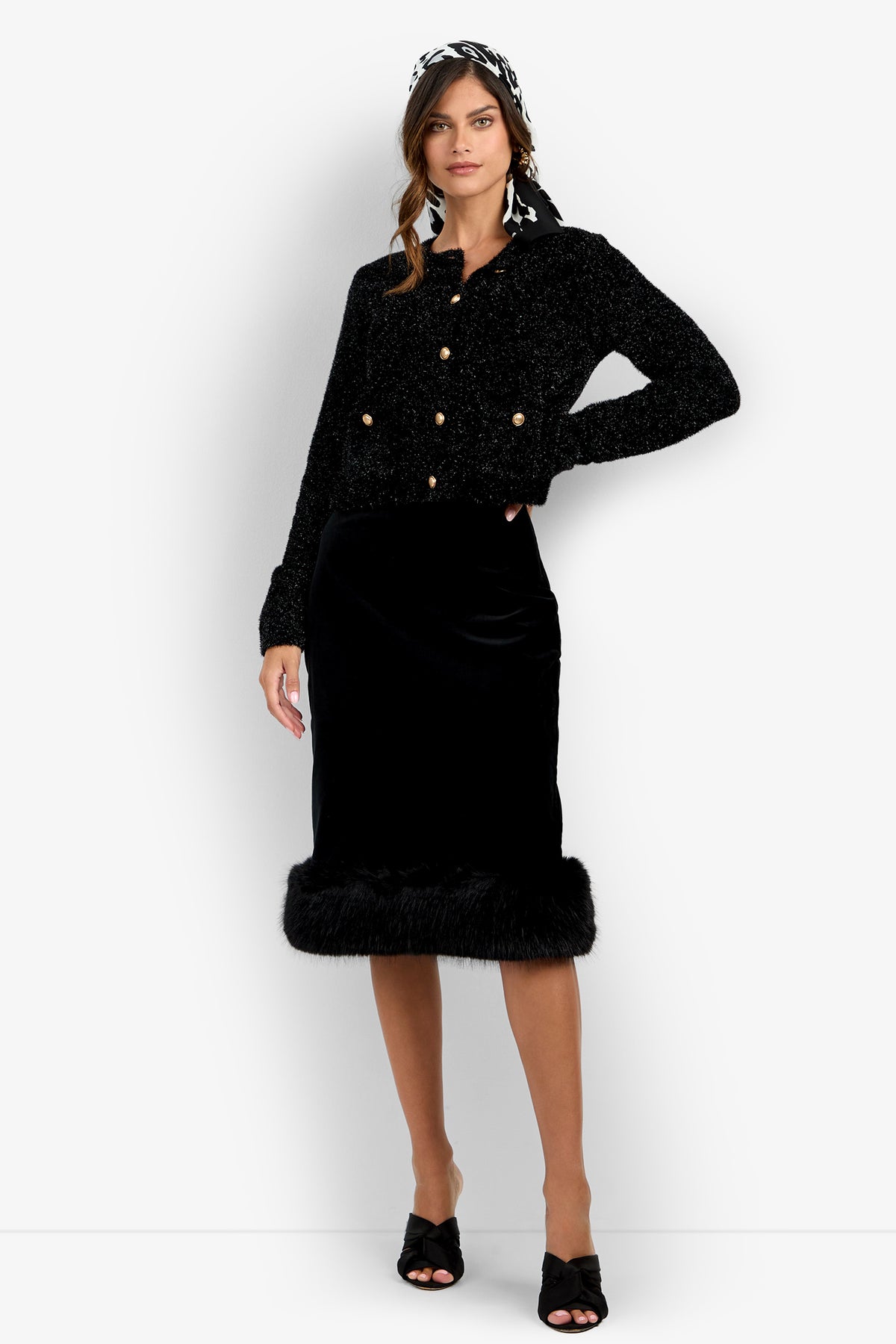 A woman wearing a black textured jacket with gold buttons, the Klara Skirt—a black velvet skirt with fur trim—black heeled sandals, and a patterned headscarf, standing against a white background.