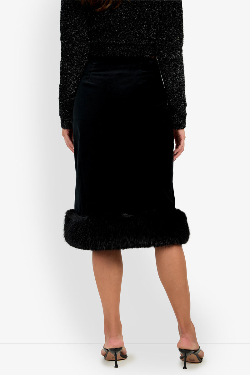 A woman stands facing away, wearing the Klara Skirt—a black midi skirt with a fur trim at the hem—paired with a sparkly black long-sleeve top and open-toe heels. Only her body from shoulders to feet is visible.