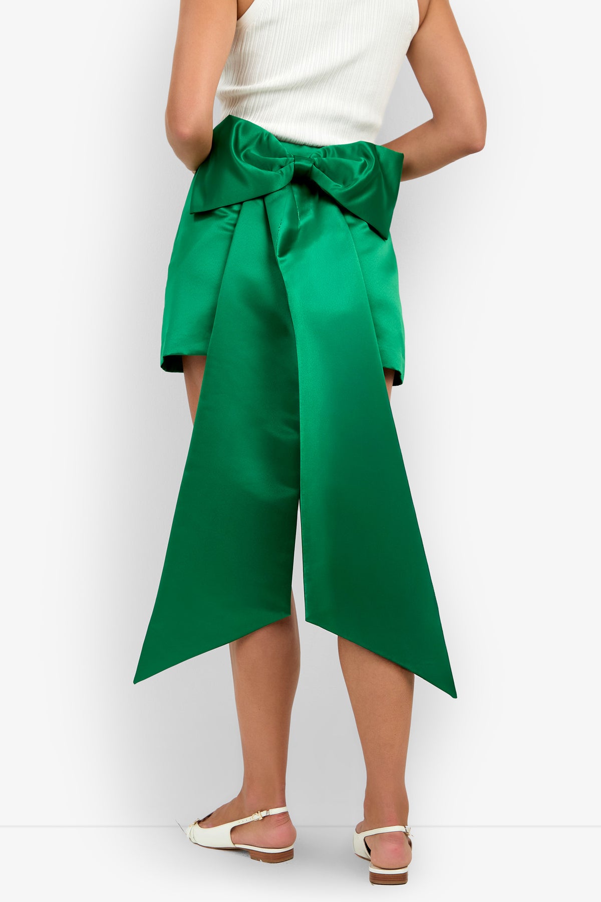 A person wearing a white sleeveless top and the Vinna Skirt—a green high-waisted skirt featuring a large, dramatic satin bow with long tails at the back—paired with white sandals.