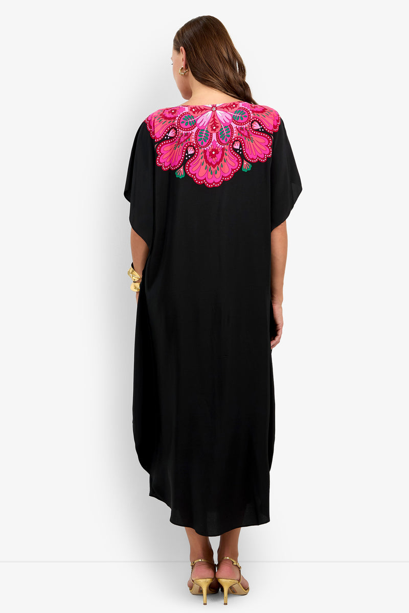 A woman with long brown hair, wearing yellow heels, is seen from behind in the Jalani Maxi Caftan Dress—a long black dress featuring vibrant pink, purple, and green embroidered floral designs on the upper back.