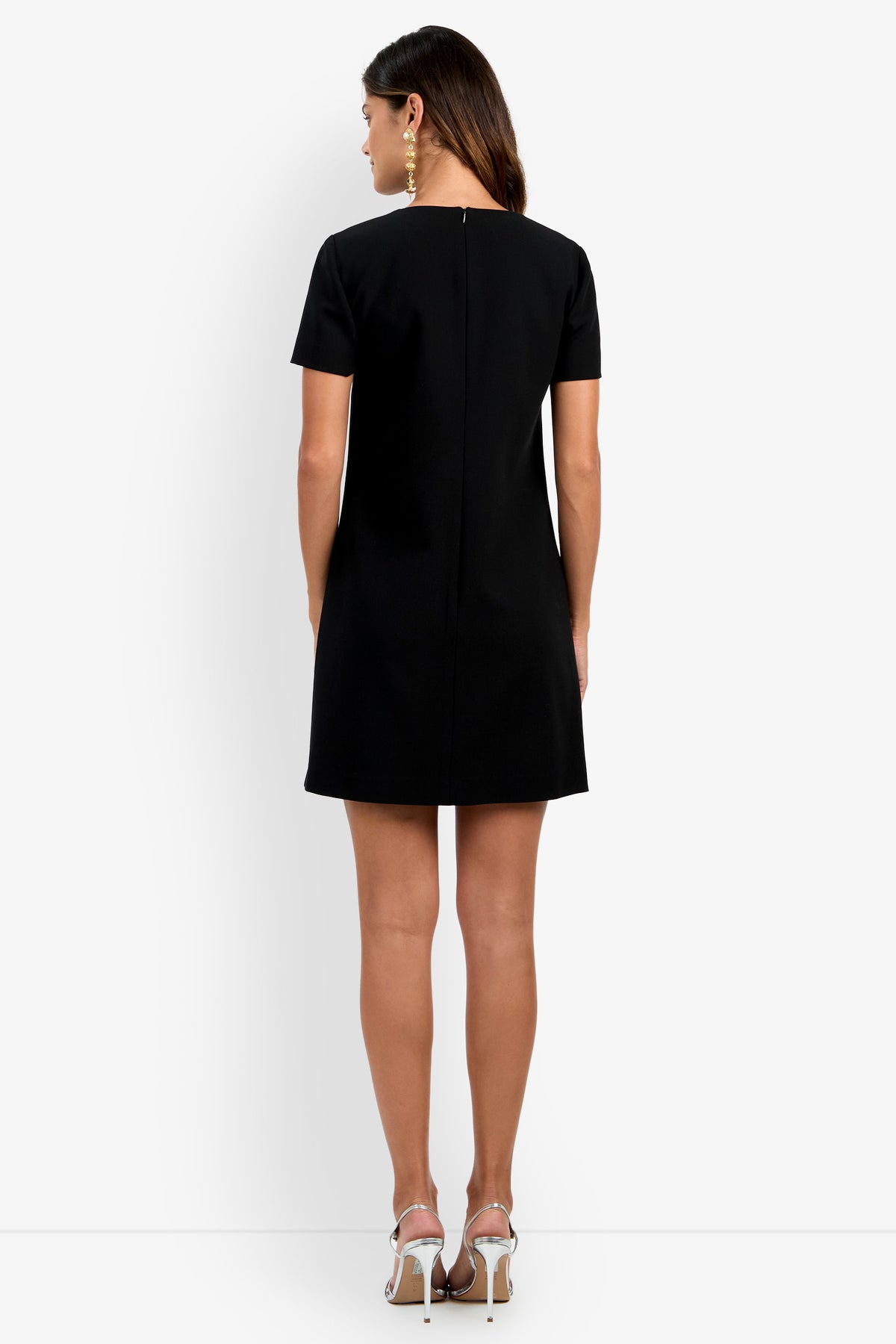 A woman stands facing away against a plain white background, wearing the Manina Dress—a short black dress with short sleeves—and white high-heeled sandals.