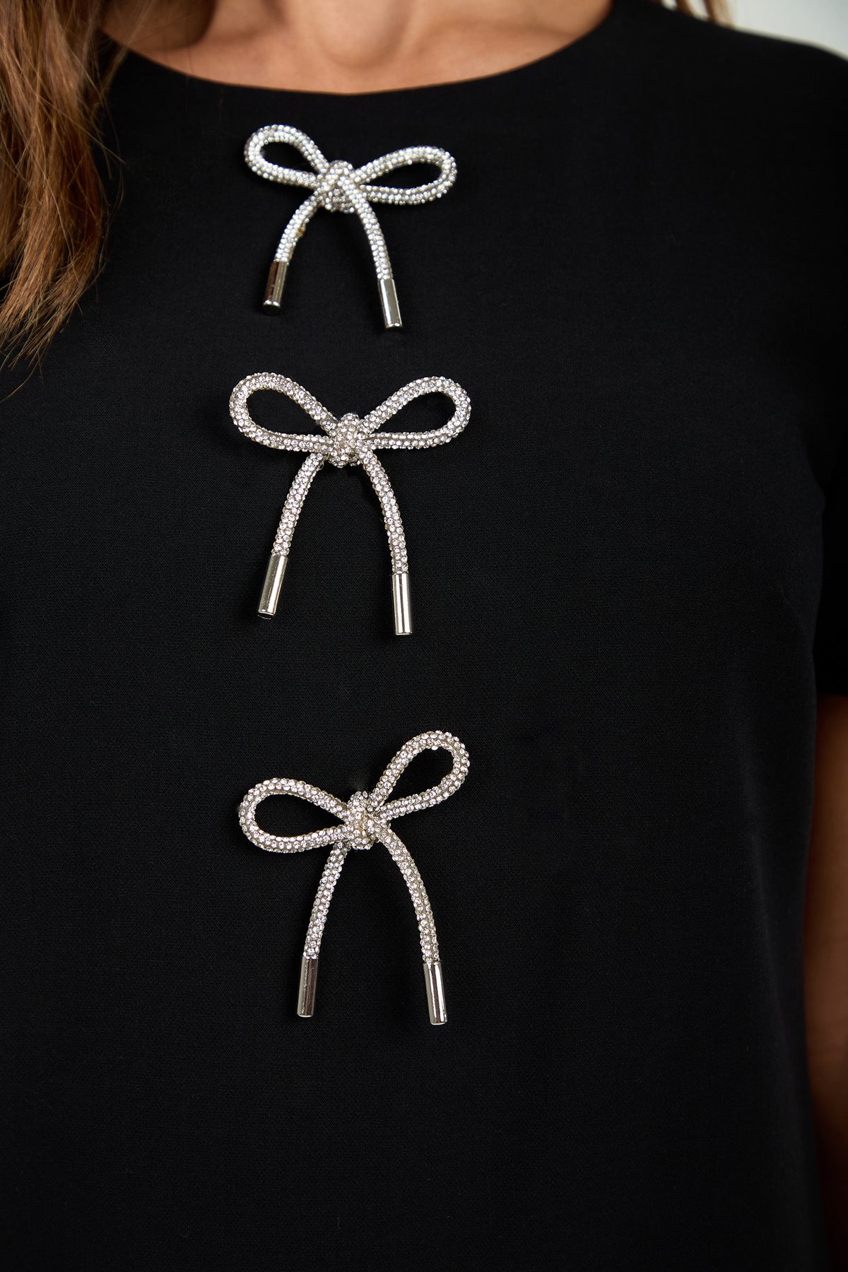Close-up of the Manina Dress in black, featuring three vertically arranged decorative silver bow-shaped embellishments on the chest area.