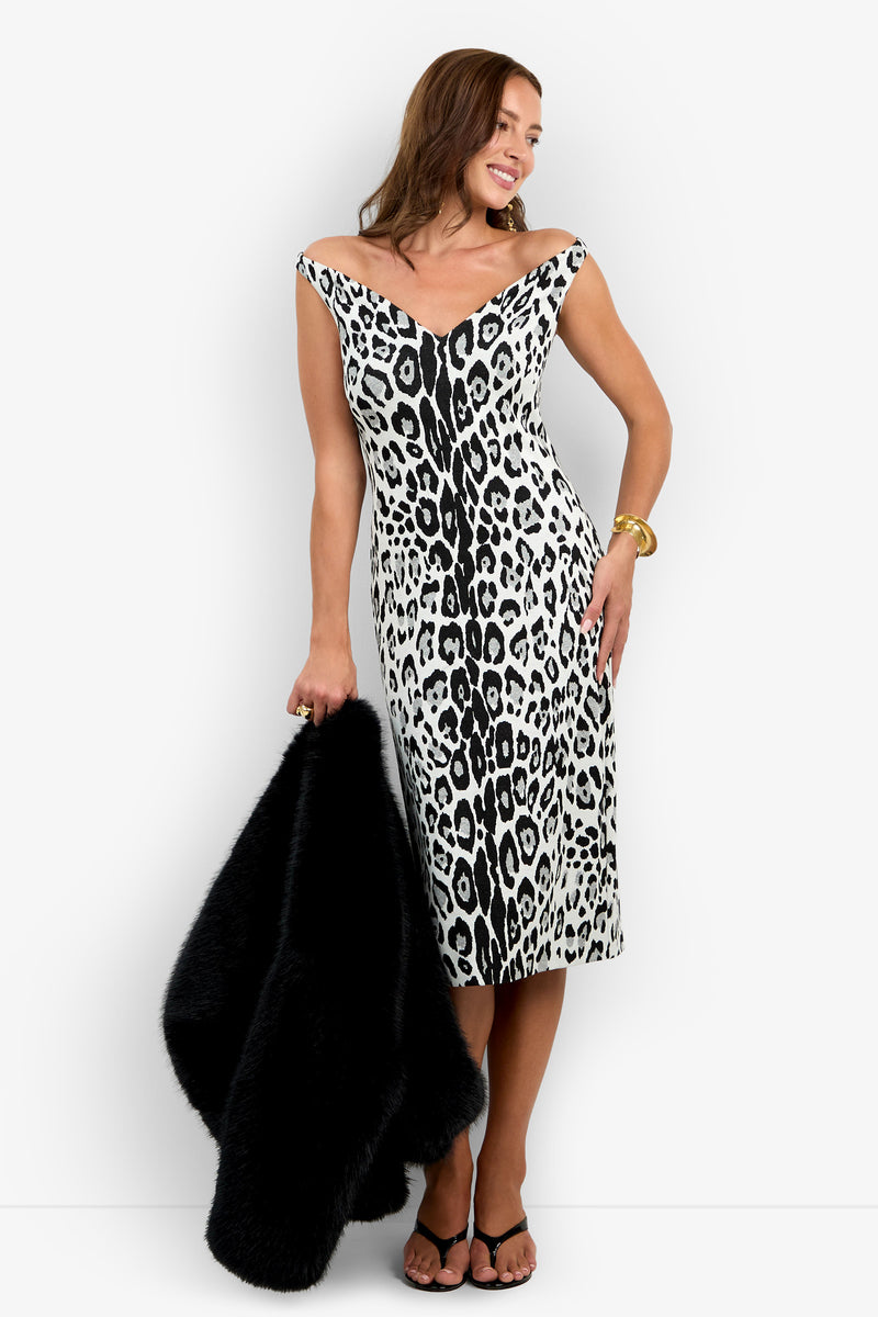 A woman wearing the Emelda Dress, an off-shoulder, black and white leopard print style, smiles as she holds a black fur coat in one hand, accessorized with gold bracelets and black sandals.