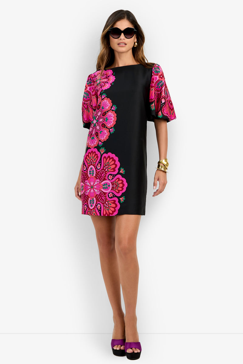 A woman stands against a white background wearing the Luv Dress—a black shift dress featuring large pink floral patterns and puffed short sleeves—paired with dark sunglasses, gold bracelets, and purple heels.
