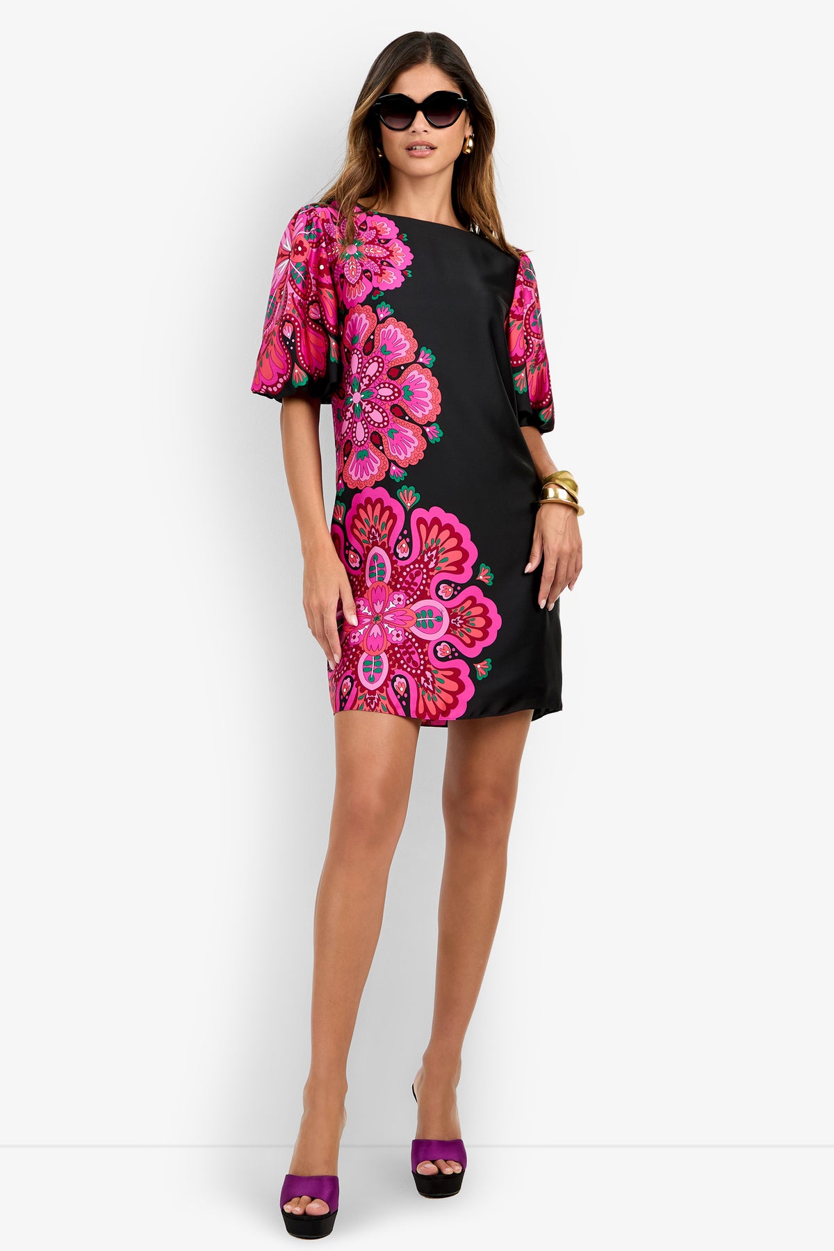 A woman wearing the Luv Dress—a short black dress with bold pink floral patterns—styled with black sunglasses, gold bangles, and purple platform heels, standing against a plain white background.