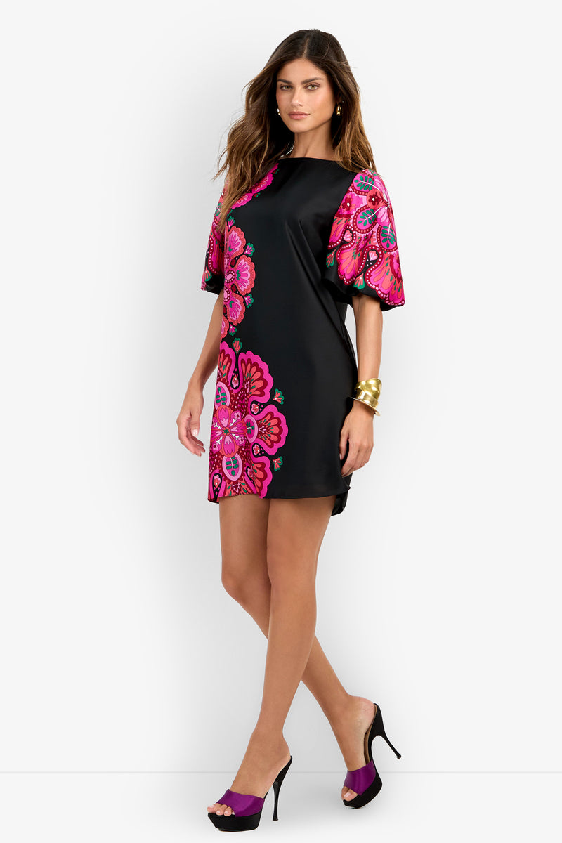 A woman models the Luv Dress—a black mini dress adorned with bright pink floral patterns and puff sleeves. She completes the look with black open-toe heels, gold bangles, and loose wavy hair, posing against a plain white background.