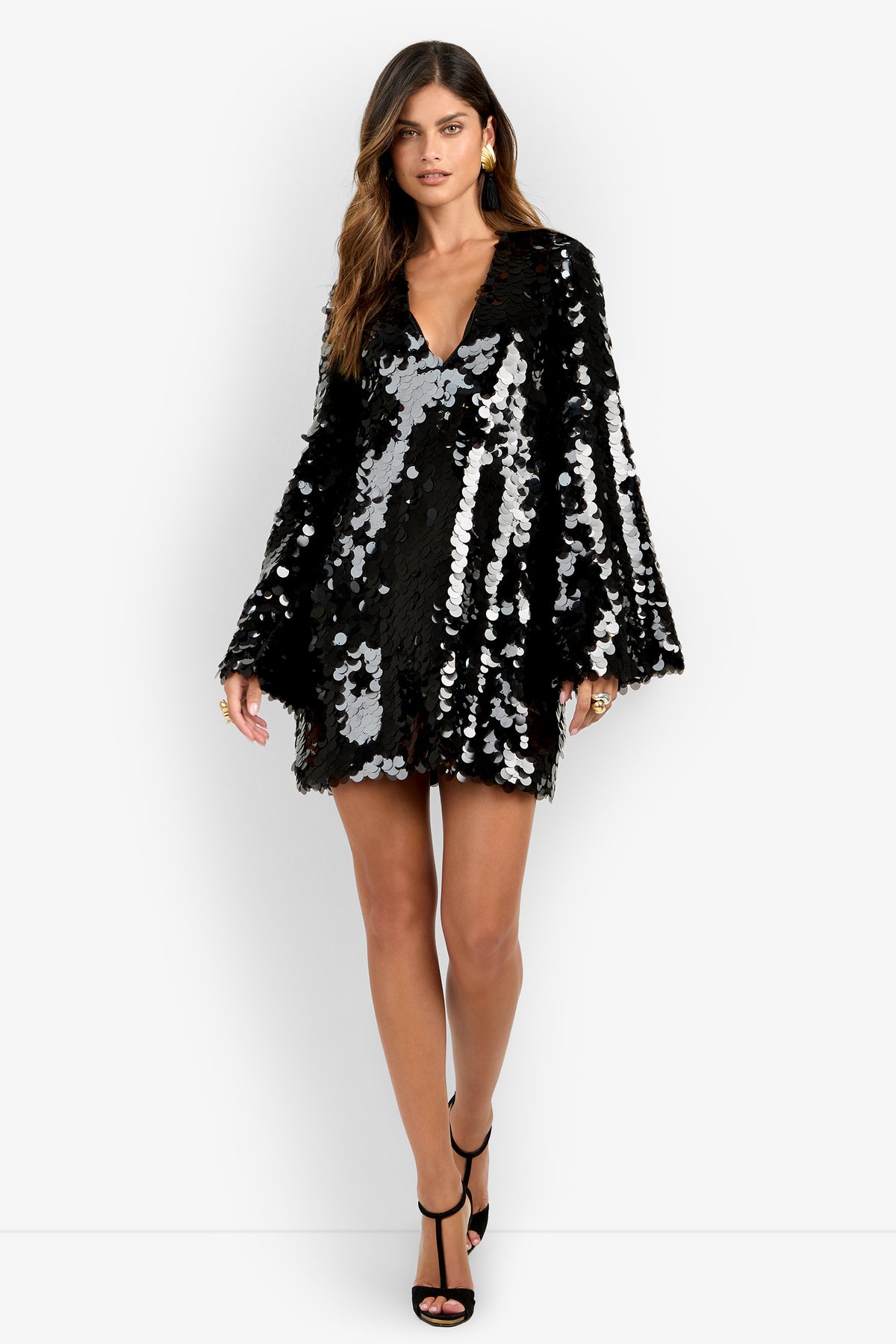 A woman models the Marea Dress, a black, long-sleeve mini dress featuring a V-neckline and bold silver sequin embellishments; she faces forward in black high-heeled sandals against a plain white background.