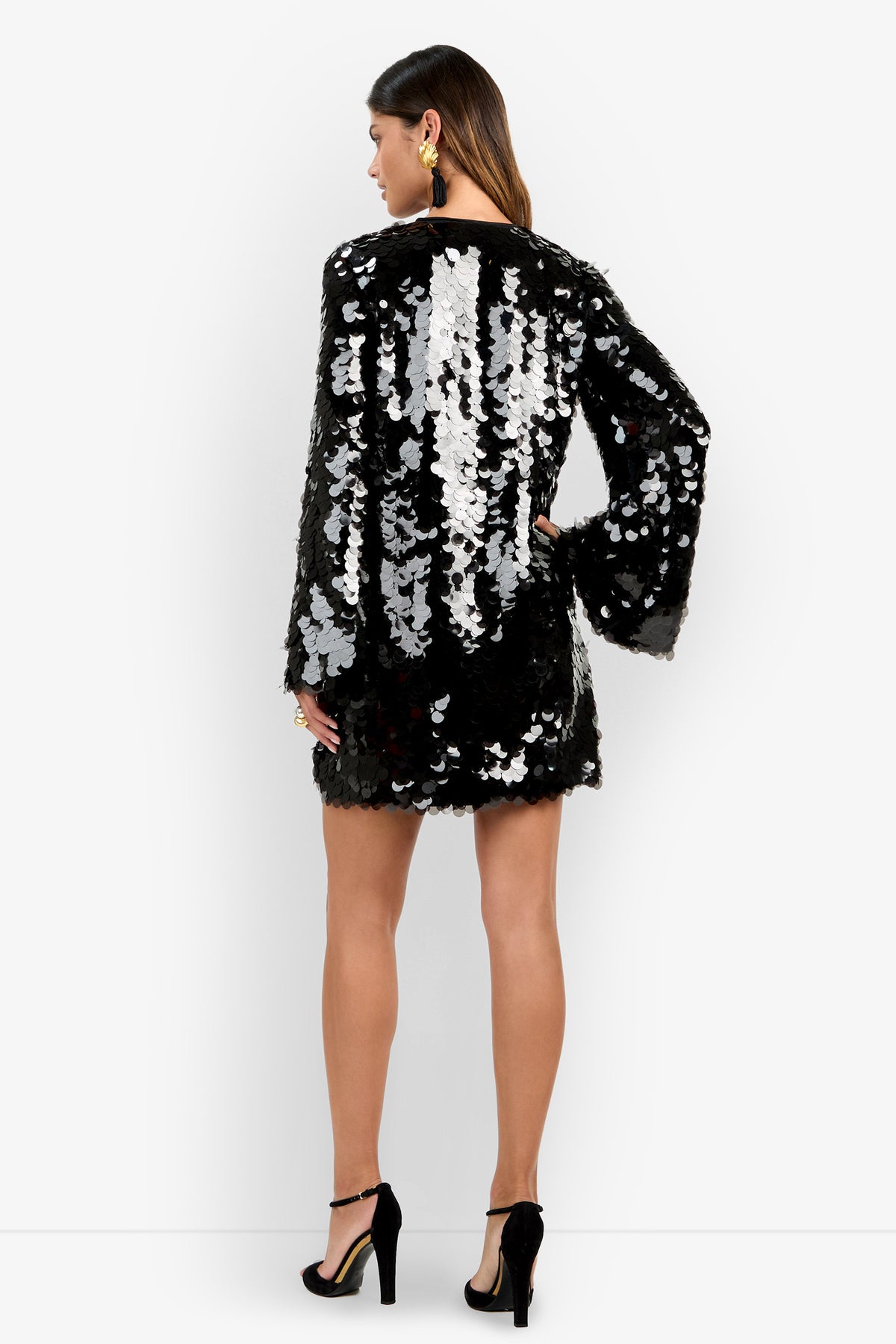 A woman stands with her back to the camera, wearing the Marea Dress—a black mini dress adorned with large silver sequins and featuring long sleeves. She pairs it with black high heels and statement earrings, set against a plain white background.