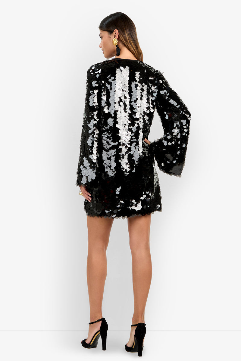 A woman stands with her back to the camera, wearing the Marea Dress—a black mini dress adorned with large silver sequins and featuring long sleeves. She pairs it with black high heels and statement earrings, set against a plain white background.