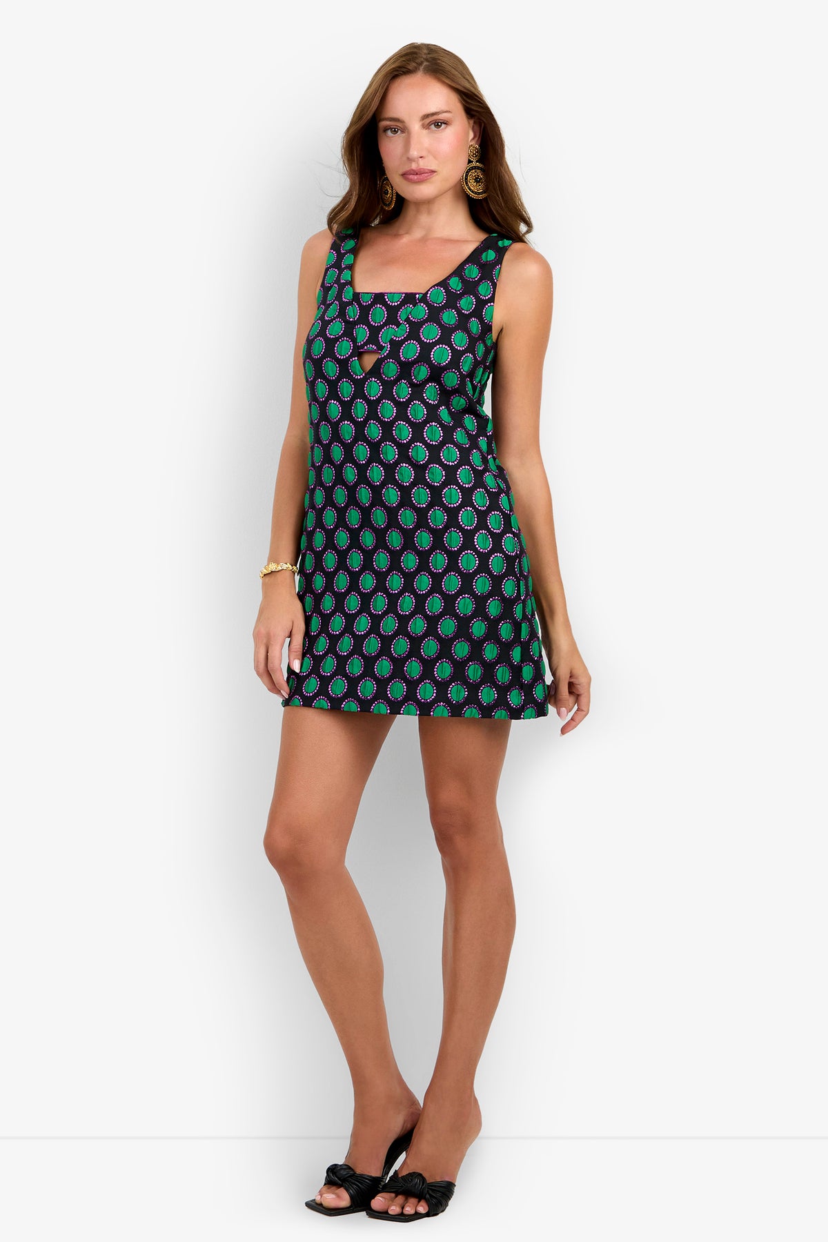 A woman wears the BAMBILLA DRESS, a sleeveless, short black dress made from soft polyester featuring a green and blue paisley pattern. She pairs it with black sandals, gold hoop earrings, and a bracelet while standing against a plain white background.