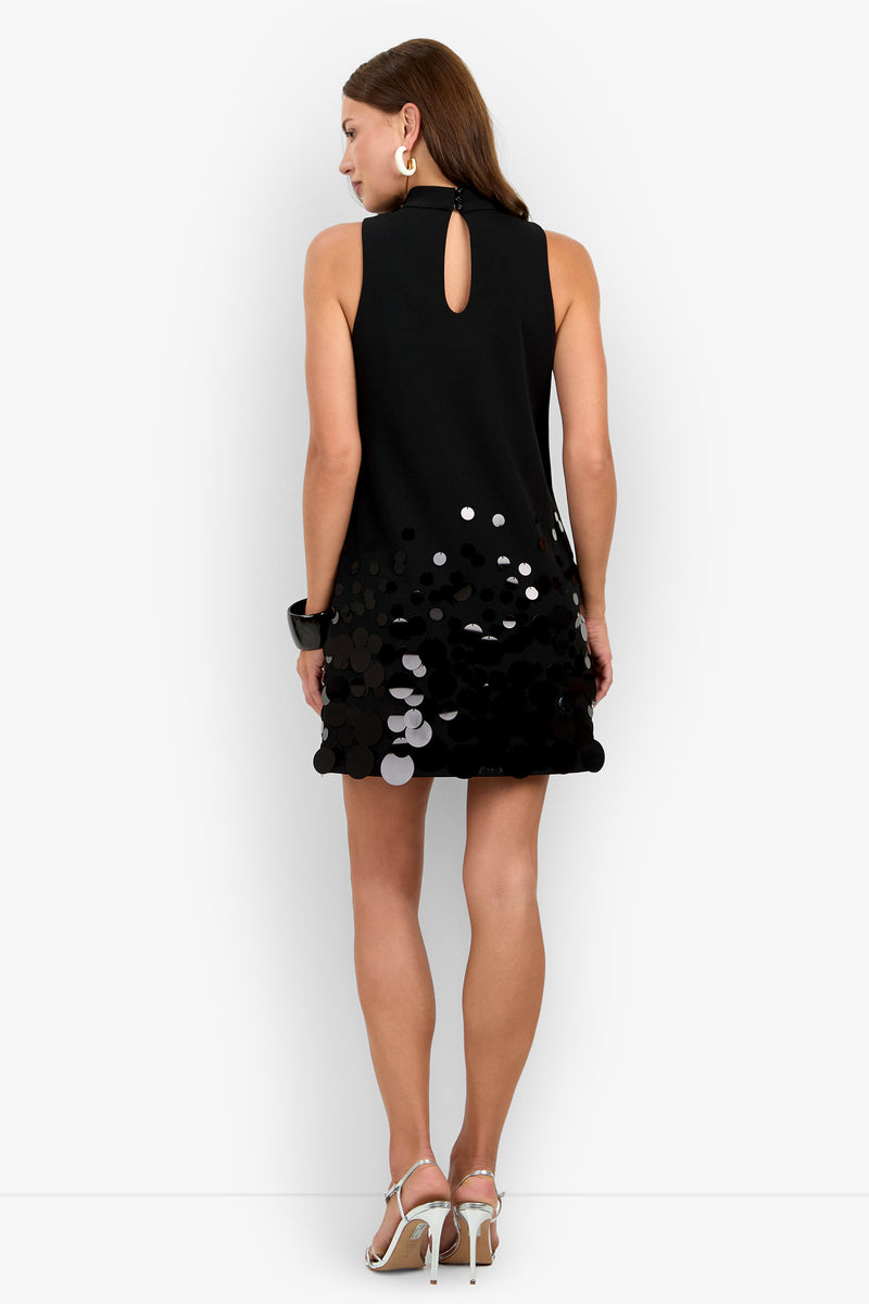 A woman stands with her back to the camera, wearing the Allena Dress—a sleeveless black dress featuring shiny sequin details at the hem—paired with silver hoop earrings, a black bracelet, and silver high-heeled sandals.
