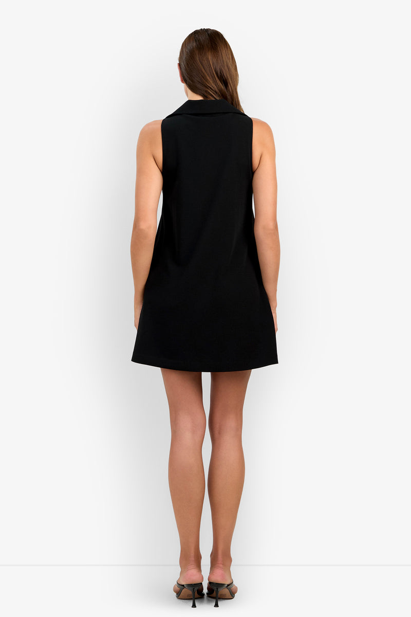 A woman with straight brown hair stands facing away against a plain white background, wearing the Siamo Dress—a sleeveless black dress—paired with black heeled sandals.