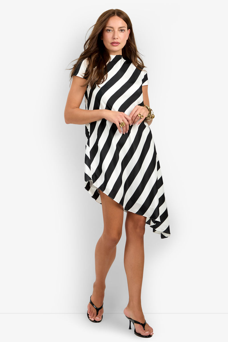 A woman wearing the Shana Dress—a black and white diagonal striped, short-sleeve dress with an asymmetrical hem—paired with black heeled sandals stands against a plain white background.