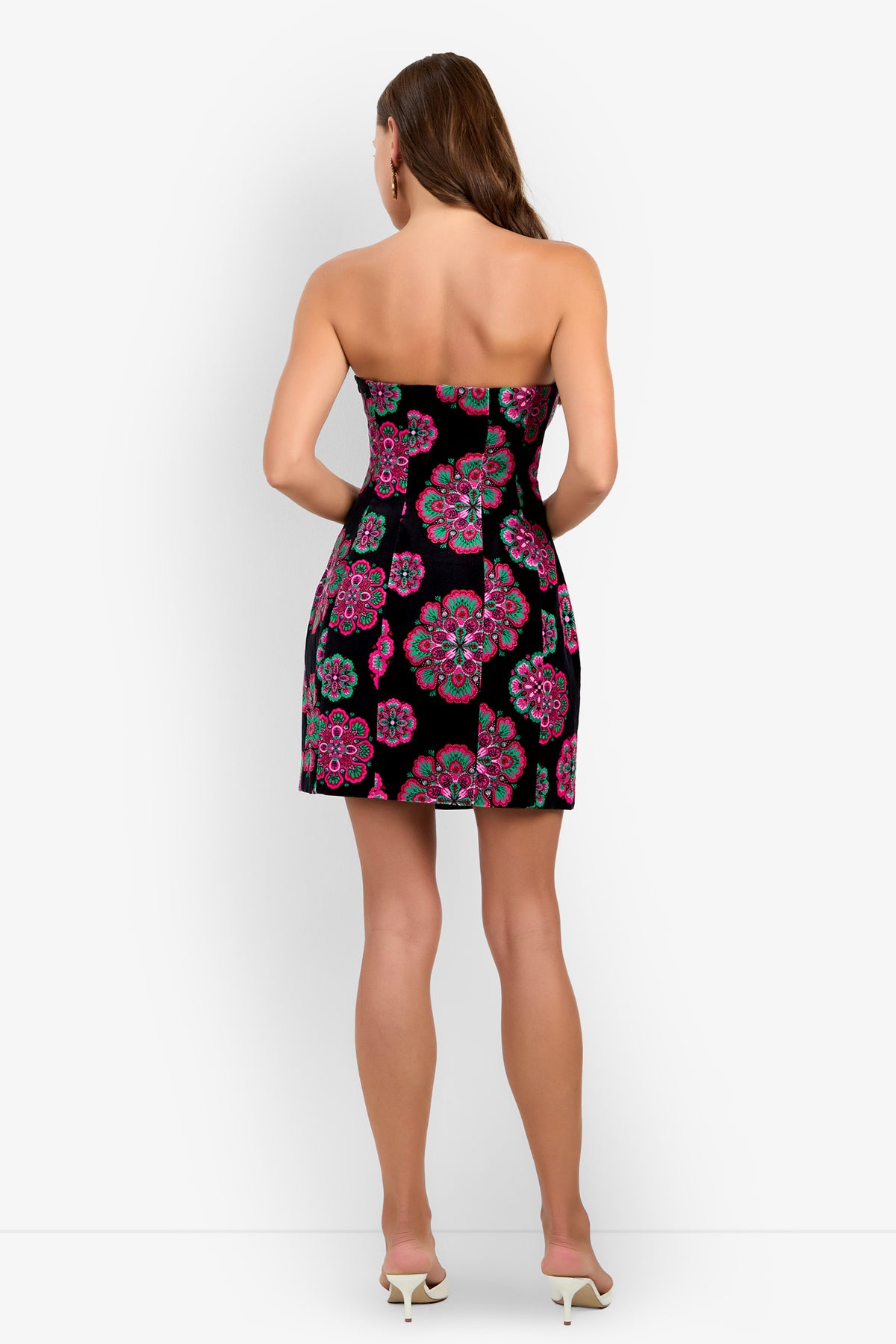 A woman stands with her back to the camera, wearing the Finale Dress—a strapless black mini dress featuring a colorful floral pattern—paired with white high-heeled shoes, set against a plain white background.