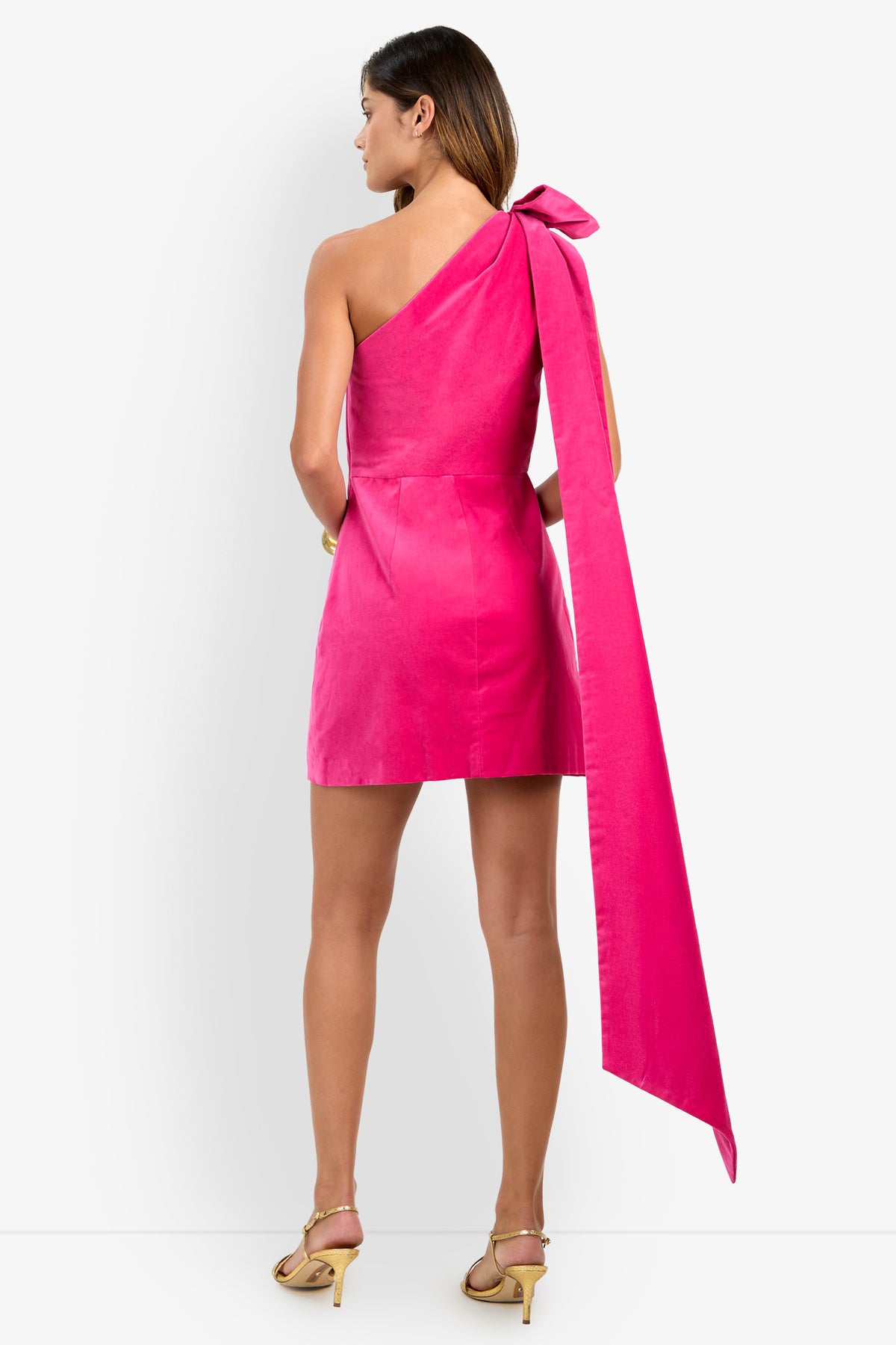 A woman stands in heels, shown from the back against a white background, wearing the Romance Dress—a bright pink, one-shoulder mini dress featuring a large bow and long sash on the shoulder.