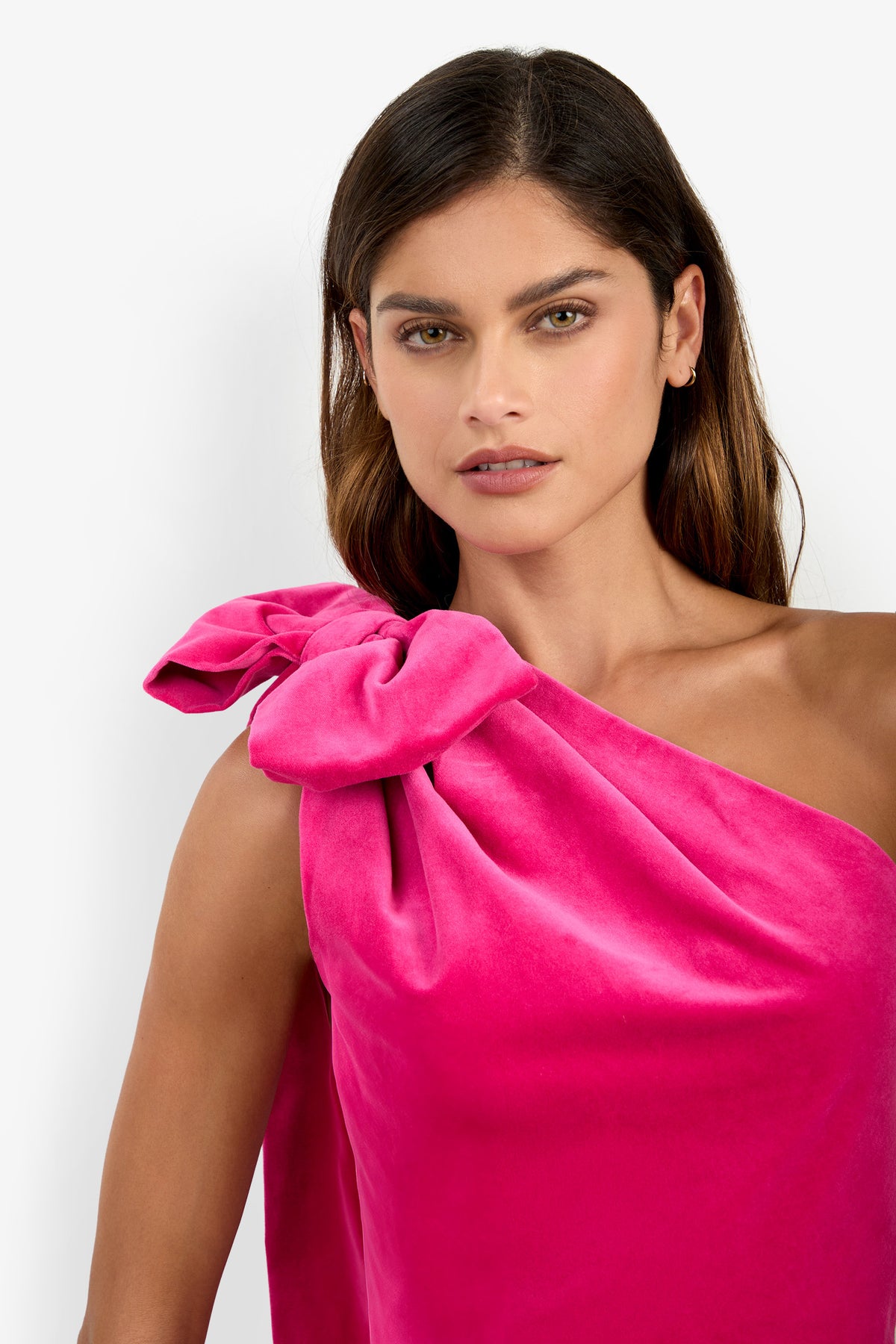 A woman with long brown hair is wearing the Romance Dress, a bright pink, one-shoulder style featuring a large bow on the left shoulder, as she stands against a plain white background.