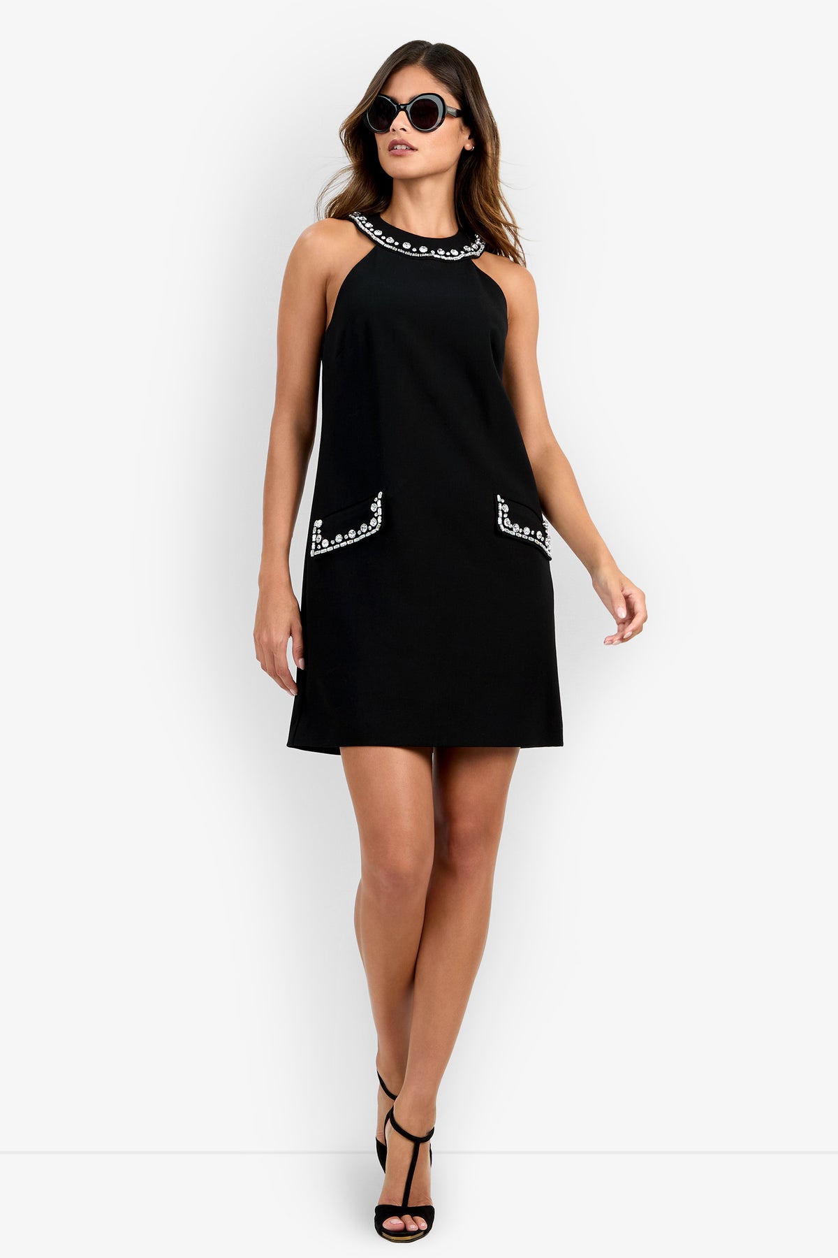 Sure! Here’s a rewritten version using your product data: A woman wearing the black Sephine Dress, featuring decorative trim on the collar and pockets, paired with black high heels and round sunglasses, stands confidently against a plain white background.