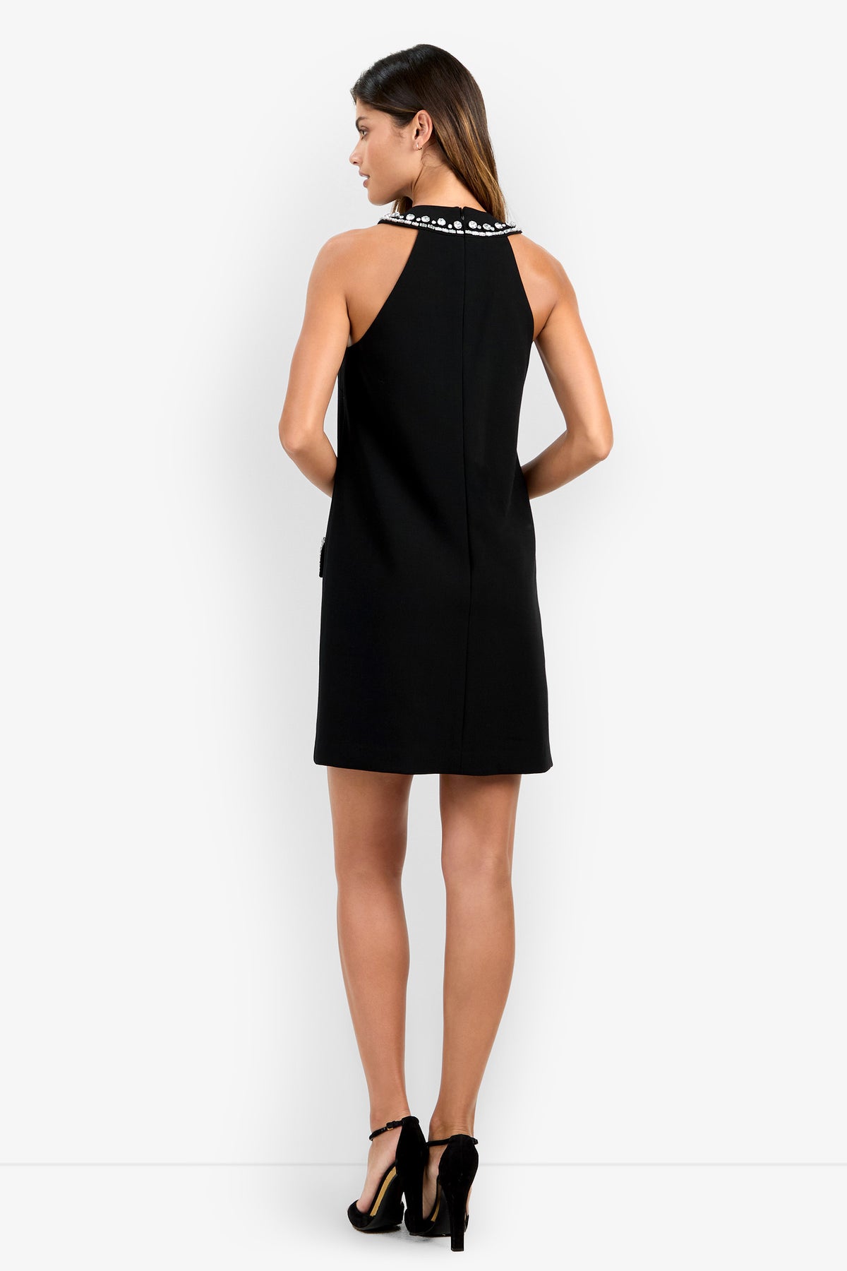 A woman with long brown hair, standing with her back to the camera against a plain white background, models the sleeveless, knee-length black Sephine Dress featuring a decorative neckline and paired with black high heels.