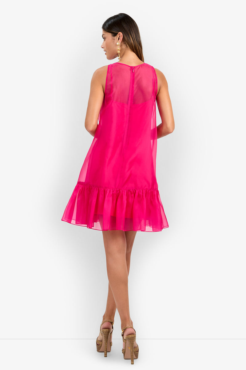 A woman is seen from behind wearing the Winner Dress—a sleeveless, bright pink dress featuring a sheer upper section and a ruffled hem. She has long hair, accessorizes with large earrings, and completes her look with high-heeled sandals.