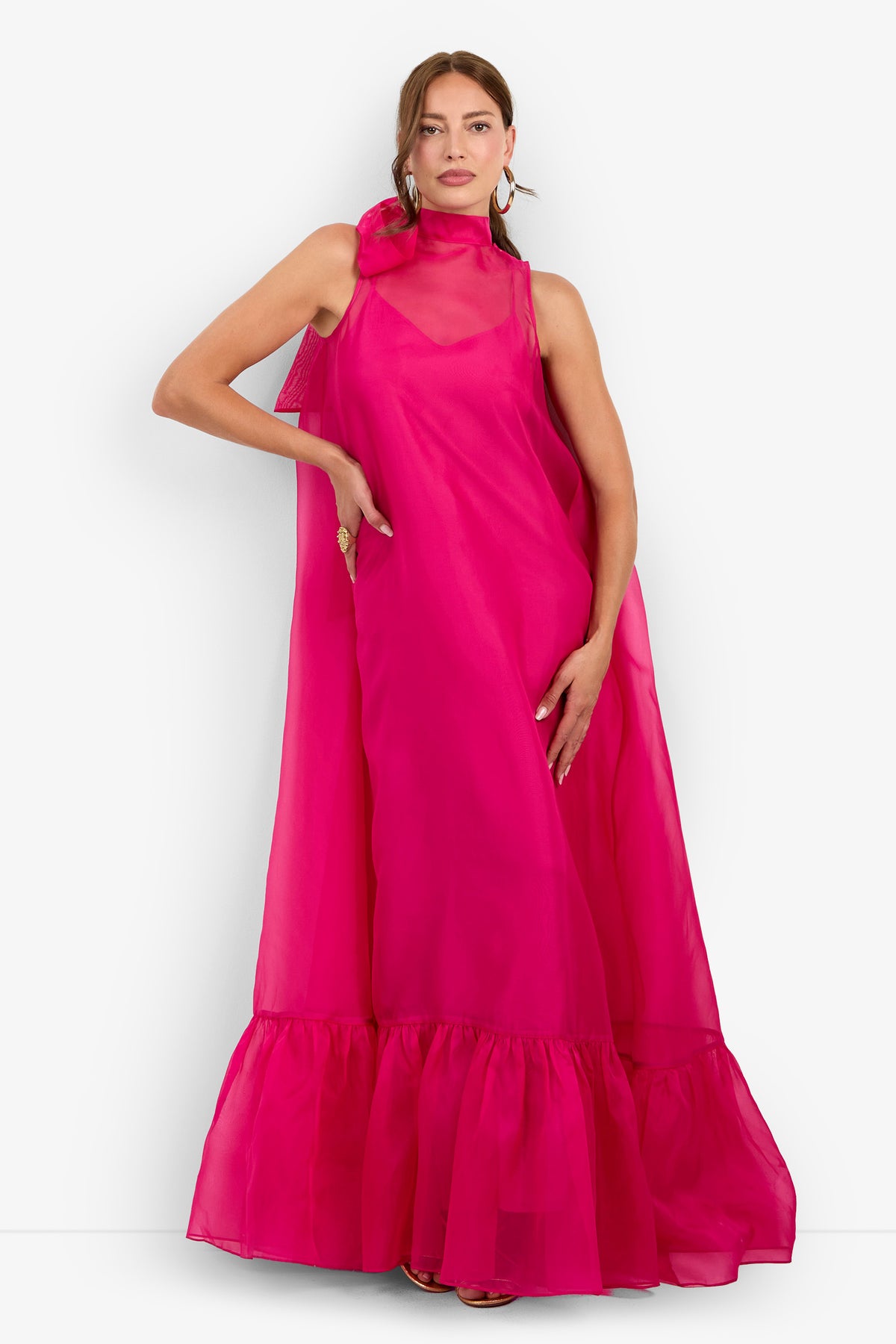 A woman wearing the Leading Dress, a sleeveless, bright pink, floor-length gown featuring a ruffled hem and a sheer high neckline, stands against a white background.