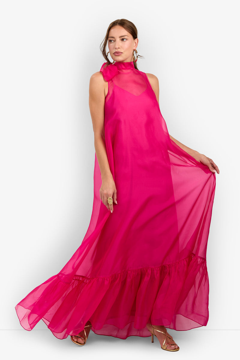 A woman stands against a plain white background wearing the Leading Dress—a vibrant, sleeveless, hot pink sheer maxi dress with a ruffled hem—paired with gold hoop earrings and strappy heels.