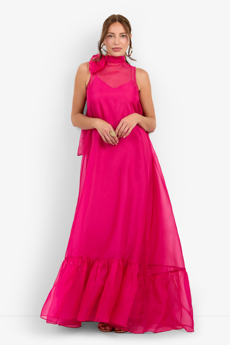 A woman stands against a white background wearing the Leading Dress in Tormalina—a vibrant, sleeveless, floor-length gown featuring a ruffled hem and a sheer high neckline.