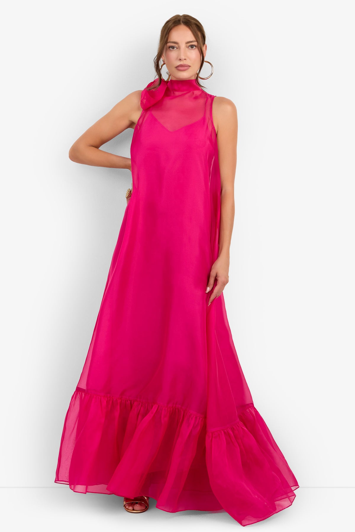 A woman wearing the Leading Dress—a sleeveless, floor-length gown in bright pink featuring a sheer high neckline and ruffled hem—poses with one hand on her hip against a white background.