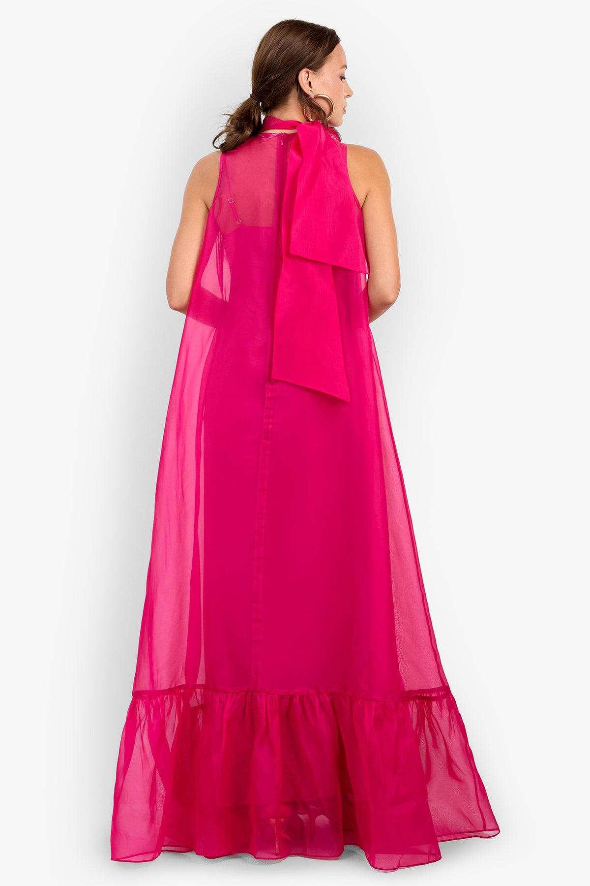 A woman is seen from behind, wearing the Leading Dress—a sleeveless, bright pink sheer maxi dress featuring a ruffled hem and an oversized bow at the neckline. The floor-length, flowy dress has a visible inner lining and is set against a plain white background.