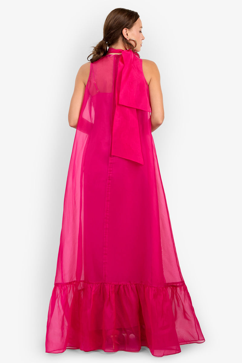 A woman is seen from behind, wearing the Leading Dress—a sleeveless, bright pink sheer maxi dress featuring a ruffled hem and an oversized bow at the neckline. The floor-length, flowy dress has a visible inner lining and is set against a plain white background.