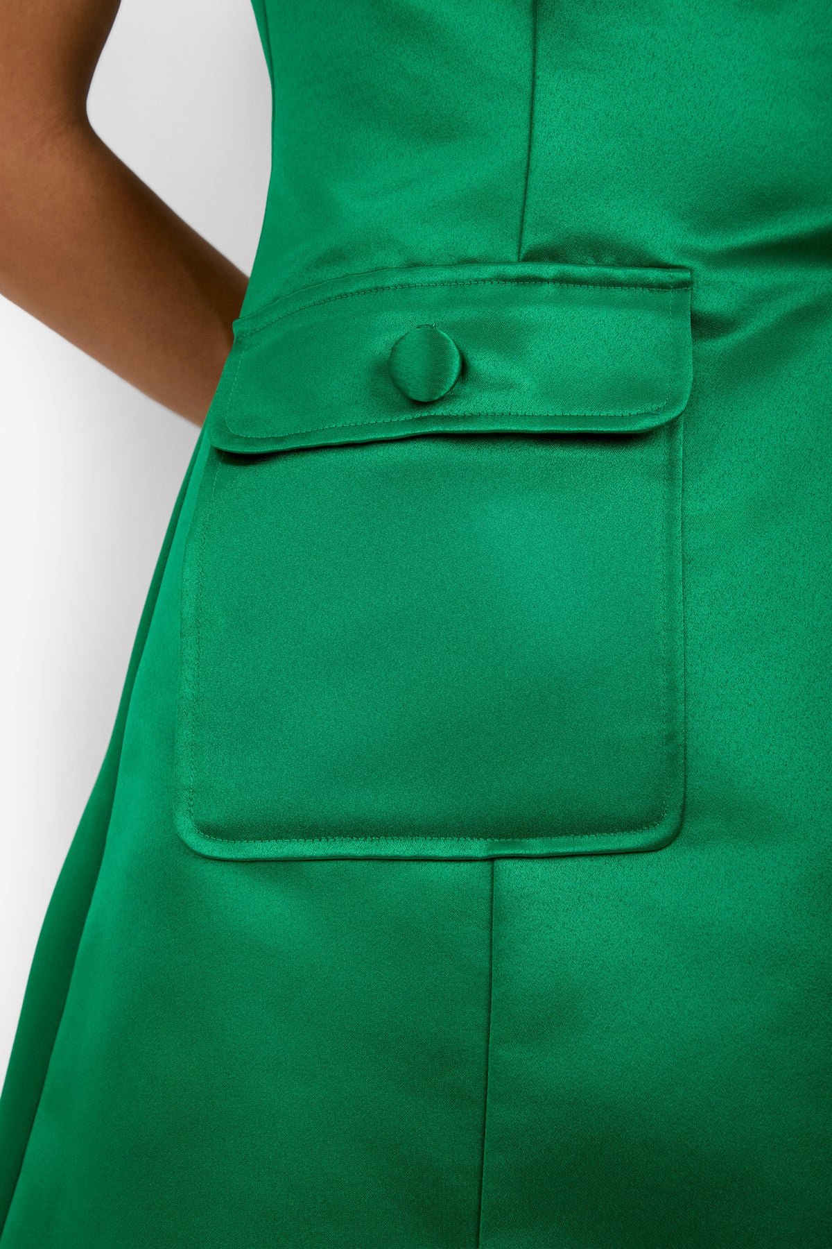 Close-up of the Guinevere Dress in green satin, showcasing a large pocket with a flap and a covered button. Only part of the wearer’s arm is visible on the left side of the image.