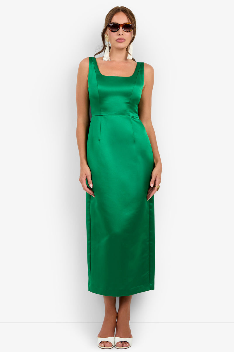 A woman wearing the Philia Dress—a sleeveless, emerald green satin dress with a square neckline—paired with white tassel earrings, sunglasses, and white heels, stands against a white background.