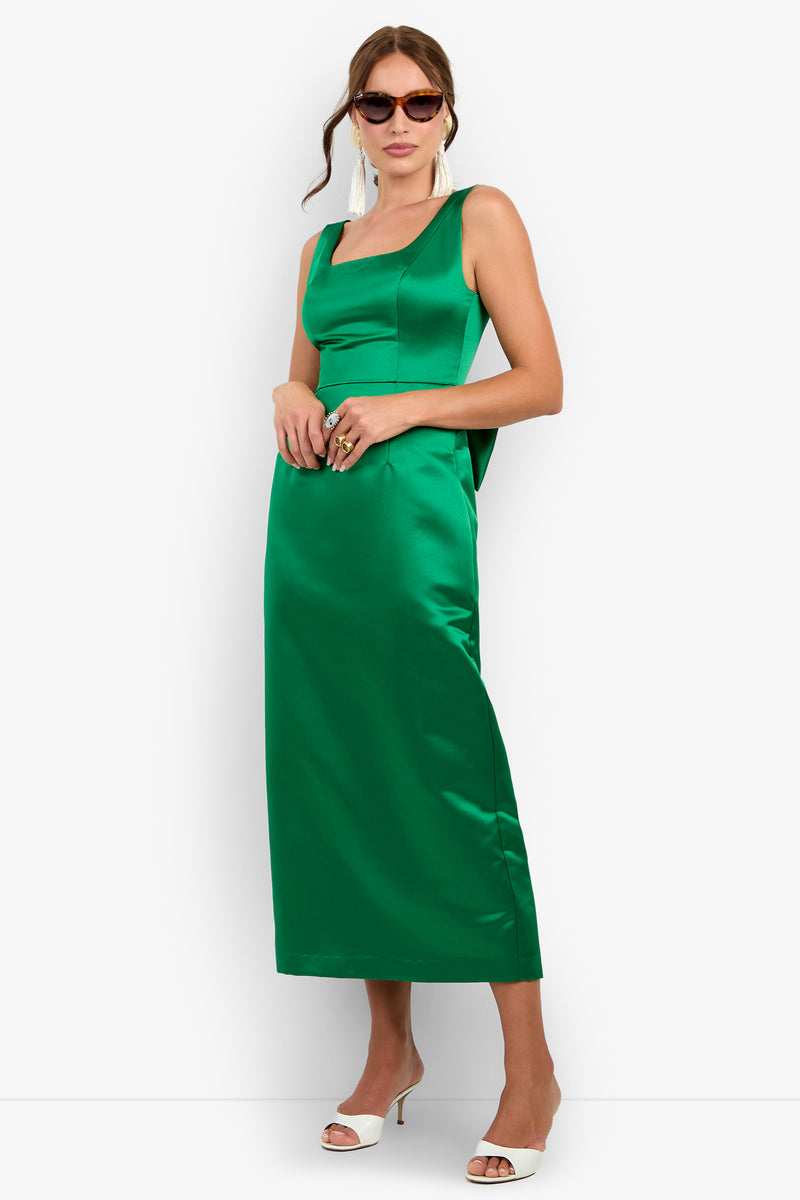 A woman wearing the Philia Dress, a sleeveless green satin design, paired with white heeled sandals, drop earrings, and brown sunglasses, stands against a plain white background.