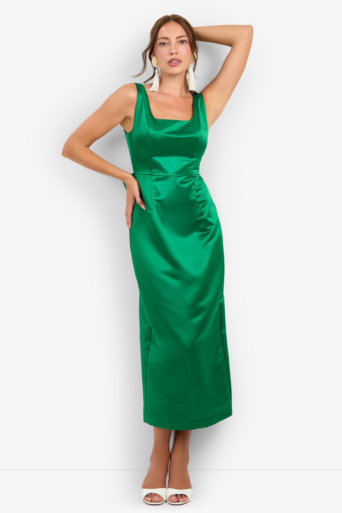 A woman wearing the Philia Dress—a sleeveless, fitted, emerald green satin dress with a square neckline—poses against a white background with one arm raised and the other on her hip, accessorizing with white earrings and white open-toed heels.