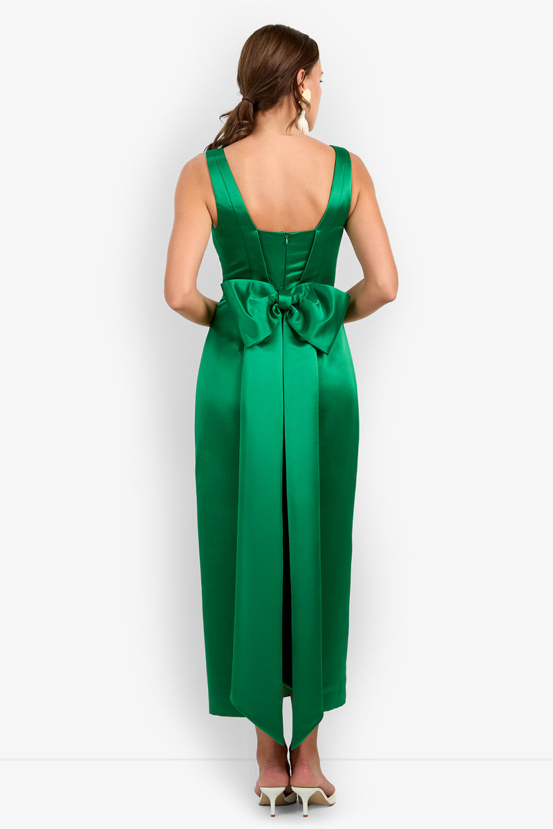 A woman stands facing away from the camera wearing the Philia Dress, a sleeveless, emerald green satin gown featuring a large bow at the lower back. She pairs the dress with white heels and has brown, shoulder-length hair.
